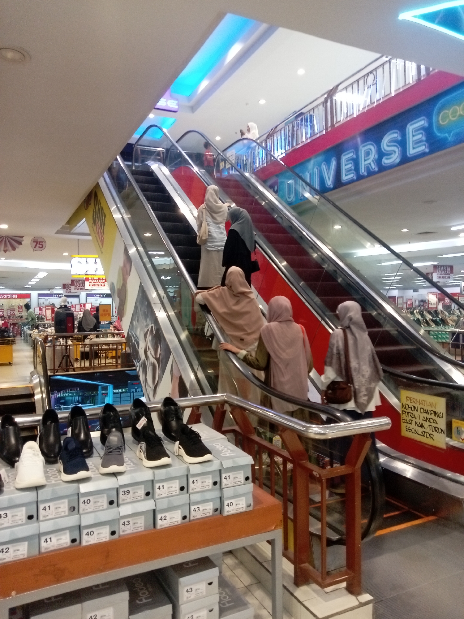 The image depicts the interior of a shopping mall in Batam, Indonesia, specifically showing an escalator area with several people ascending and descending. The mall features brightly lit signage, including a large blue sign that reads "UNIVERSE COCO". On the foreground, a display of shoeboxes and shoes is visible, with various sizes indicated on the boxes. The people in the image are mostly women wearing hijabs, engaged in the act of using the escalators. The environment appears to be a commercial space with multiple levels, suggesting a busy retail setting.