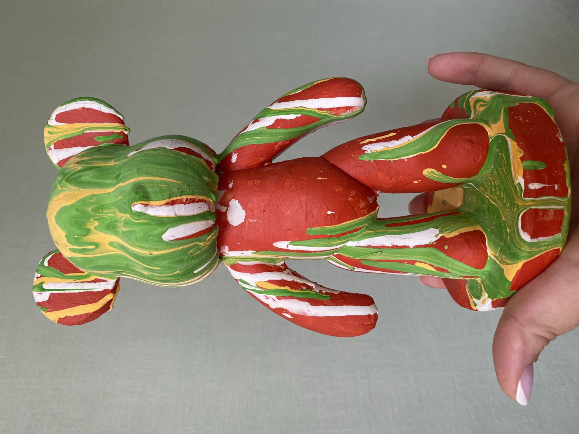 A person's hand is holding a painted, stuffed bear toy against a plain, light-colored background. The toy is primarily colored with red and green paint, swirled together, with some stripes of white and yellow. The bear has a round head with two smaller round ears, four limbs, and a round torso. The hand holding the toy is visible from the wrist up, and the thumbnail has a white tip. The setting appears to be indoors, possibly a studio or home. There is no contextual information to associate with the city of Sochi, Russia.