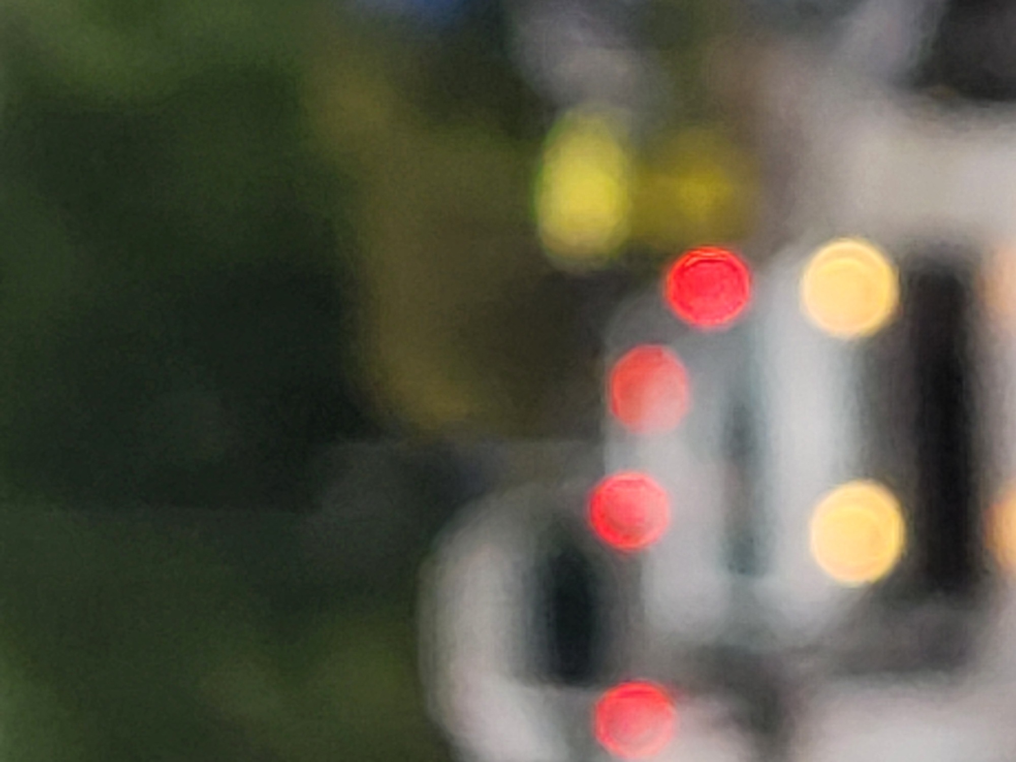 The image appears to be an out-of-focus photograph taken in an urban setting, likely Budva, Montenegro, given the prompt's reference. The left portion of the frame shows a dark green area, likely foliage. The right portion contains blurred shapes that appear to be a vehicle with red and yellow lights, possibly taillights and headlights, suggesting it is night. Details are indiscernible due to the extreme blur.