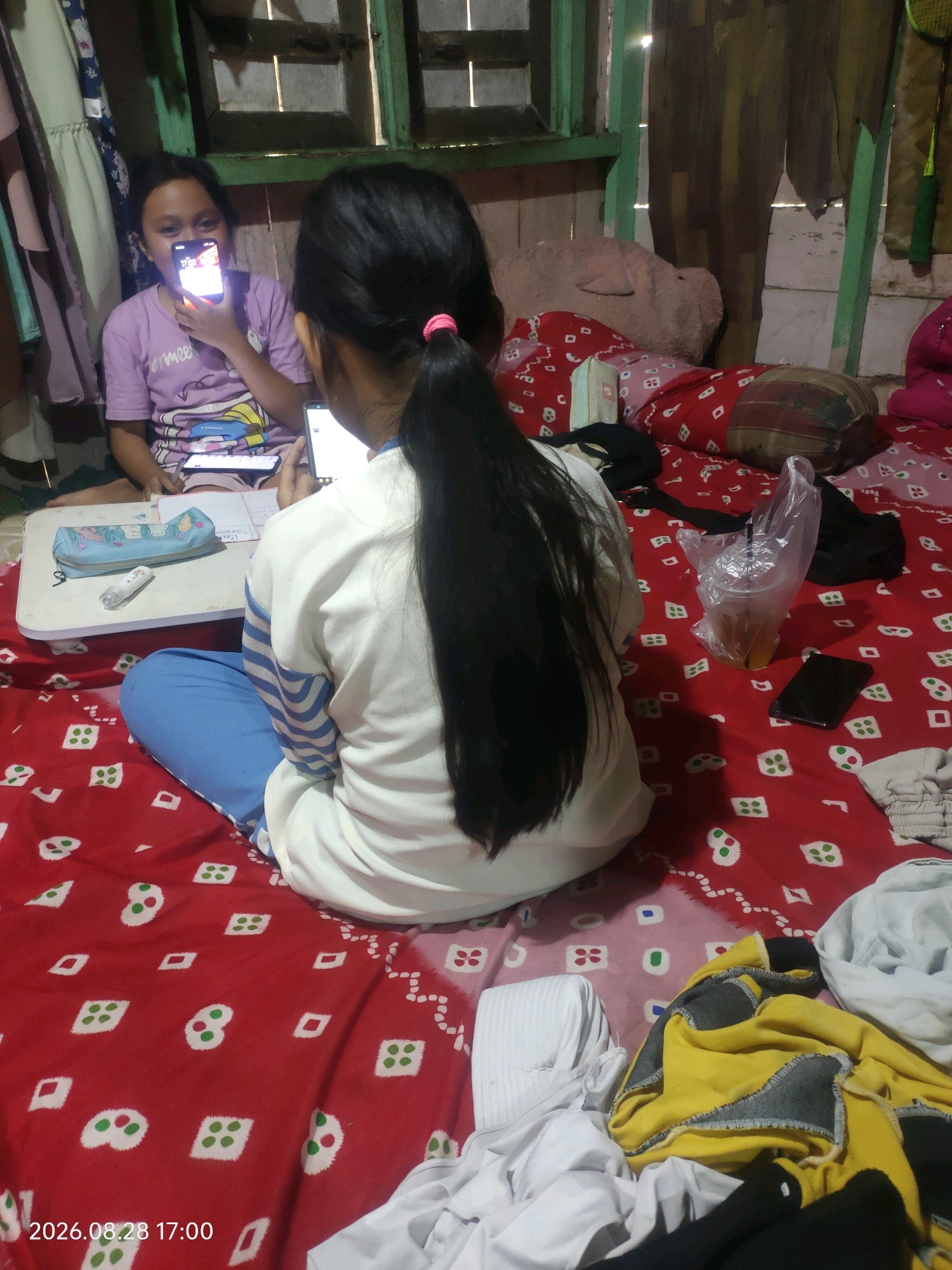 The image depicts an indoor scene, possibly a bedroom, with two young girls engaged with their mobile phones. The girl in the foreground, facing away from the camera, has long dark hair pulled back in a ponytail and wears a white and blue long-sleeved shirt and blue pants. She is holding a smartphone. To her left, another girl in a purple t-shirt is looking at her phone, which is held up and illuminated. They are seated on a red blanket with a white pattern. A small white table is in front of them, holding a blue pencil case and a bottle of liquid in a plastic bag with a straw. The background shows a wooden wall, a large pink stuffed animal, and a window with green framing. Various items of clothing, including a yellow and grey garment, are scattered on the bed. The lighting suggests it may be daytime, with some light entering from the window. A timestamp "2026.08.28 17:00" is visible in the bottom left corner. The overall setting appears to be a modest domestic environment.