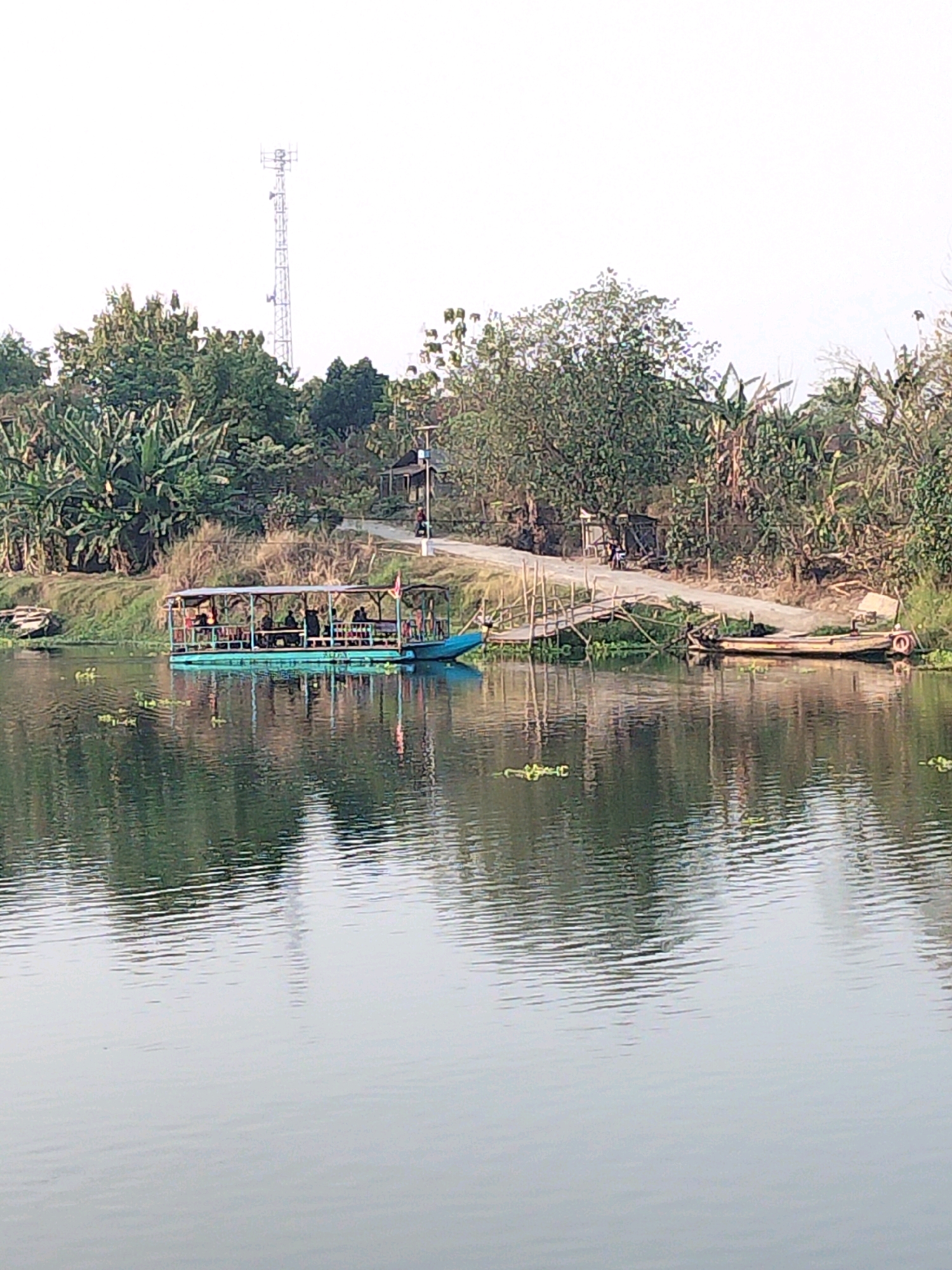 The image depicts a tranquil scene on a body of water, likely a river or canal, in Betro, Indonesia. In the foreground, the calm water reflects the surrounding environment, with ripples indicating a gentle breeze. A long, turquoise passenger boat, possibly for a tour or local transport, is moored near the bank, with several people seated under its canopy. A wooden walkway and a dirt path lead to the boat from the shore, which is lined with lush vegetation, including banana trees and other tropical plants. Further inland, a communication tower stands tall against a pale sky, suggesting the presence of modern infrastructure amidst the natural landscape. Another smaller, traditional wooden boat is also visible, tied to the bank near the walkway. The overall atmosphere is one of peaceful coexistence between nature and human activity.