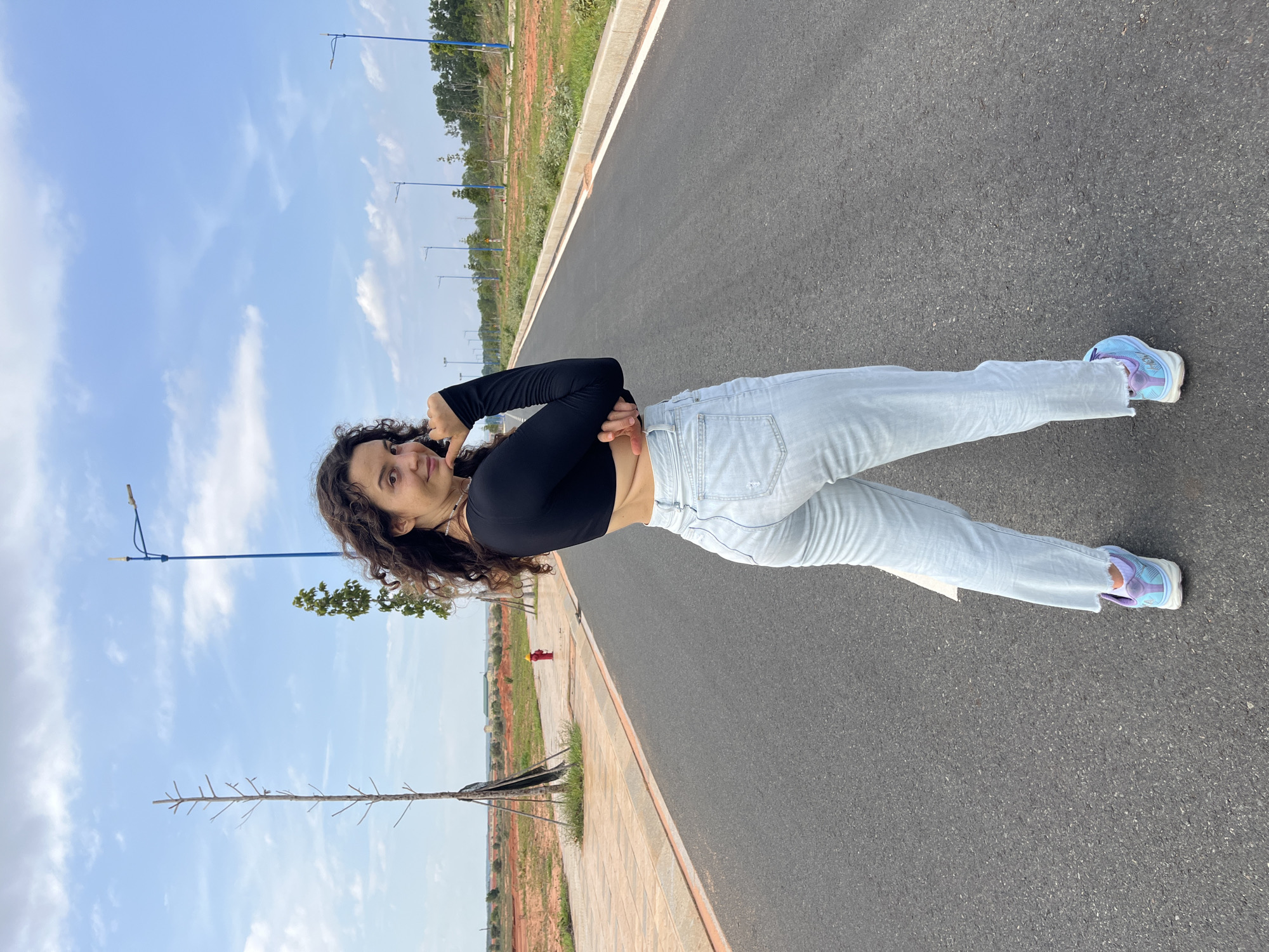 In Mui Ne, Vietnam, a young woman with curly dark hair stands on a paved road, looking over her left shoulder at the camera. She is wearing a black long-sleeved crop top and light-wash, high-waisted jeans. Her right arm is bent, with her hand touching her chin, while her left arm is crossed in front of her body. She is wearing light blue and purple athletic shoes. The road stretches into the distance, lined with sparse trees and streetlights. The landscape to the left of the road features red soil and some construction, while the right side has a grassy verge and more trees. The sky is blue with scattered white clouds.