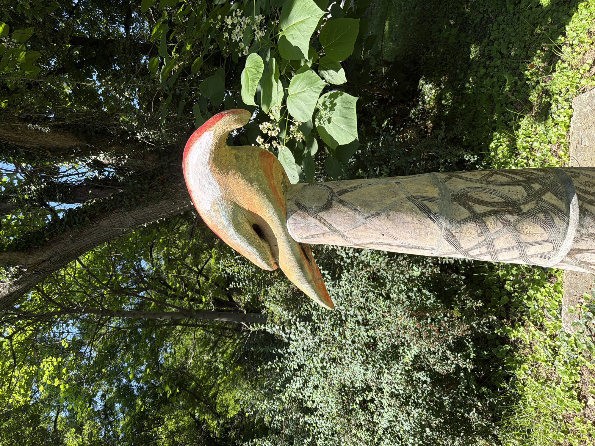 This is a close-up, eye-level outdoor shot of a colorful, abstract bird sculpture perched atop a rough-hewn, cylindrical pedestal. The sculpture is predominantly painted in shades of orange, yellow, and white, with a prominent red stripe along its curved head. The pedestal is made of a light-colored material, possibly stone or concrete, and is adorned with dark, intersecting line patterns that resemble tribal markings.
The sculpture and pedestal are set against a backdrop of lush green foliage, including trees and bushes, suggesting a garden or park setting. Sunlight filters through the leaves, creating dappled shadows on the ground. A flowering tree with small white blossoms is visible to the right of the sculpture.
The sculpture appears to be a permanent art installation. No people are present in the image. The environment is serene and natural, with the vibrant sculpture providing a focal point. The image was taken in the city of Pazardzhik, Bulgaria, and is located in the park near the Historical Museum and the Ensemble "Zornitsa".