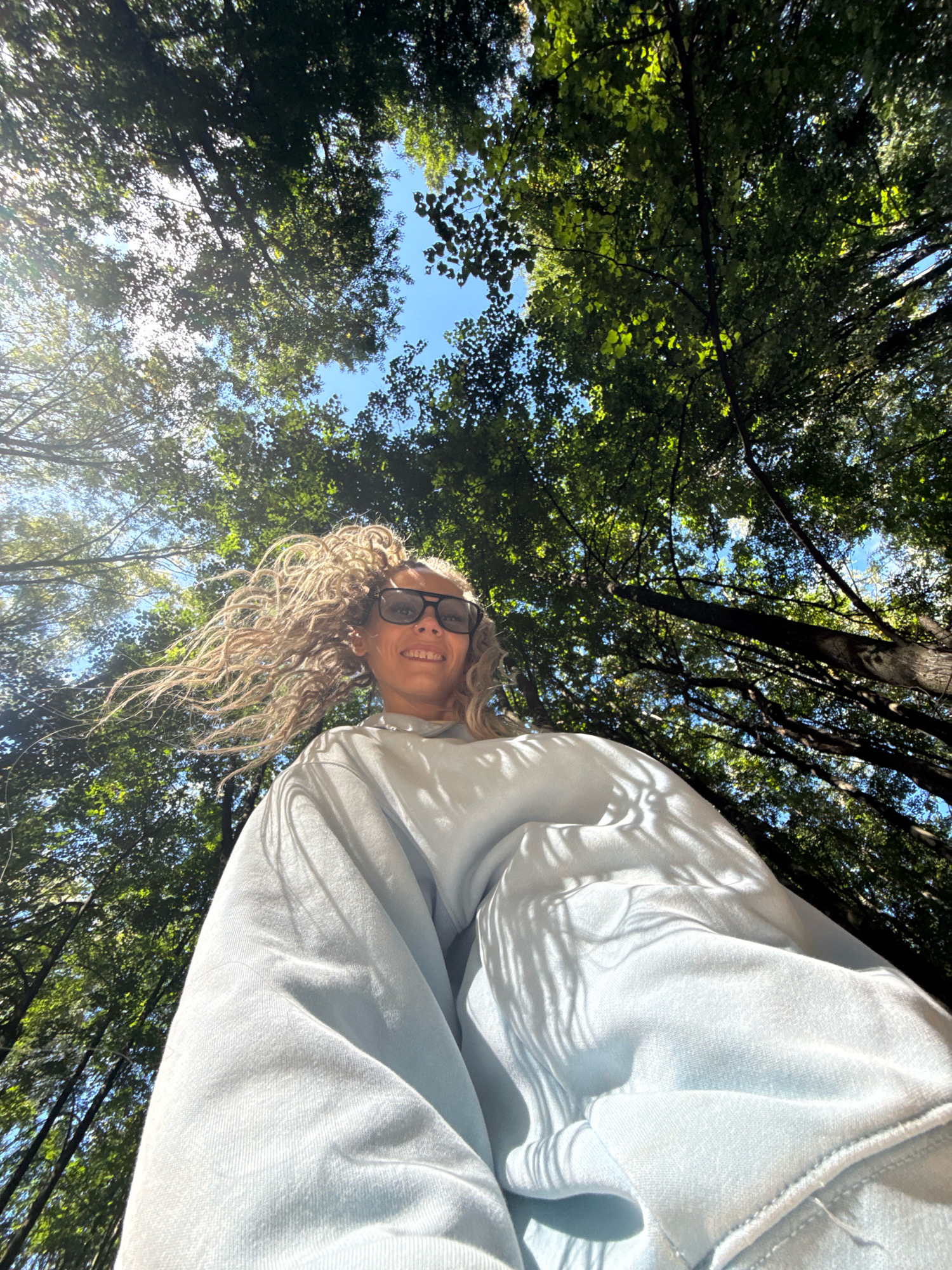 In the city of Dumino, Russia, a low-angle shot captures a woman with blonde dreadlocks and sunglasses, wearing a light blue sweatshirt, standing outdoors. She is looking upwards, smiling, with her hair blowing in the wind. The background is dominated by lush green trees and a clear blue sky, suggesting a forest or park setting. Sunlight filters through the leaves, casting shadows on the woman's sweatshirt. The perspective emphasizes the height of the trees and the vastness of the natural environment.