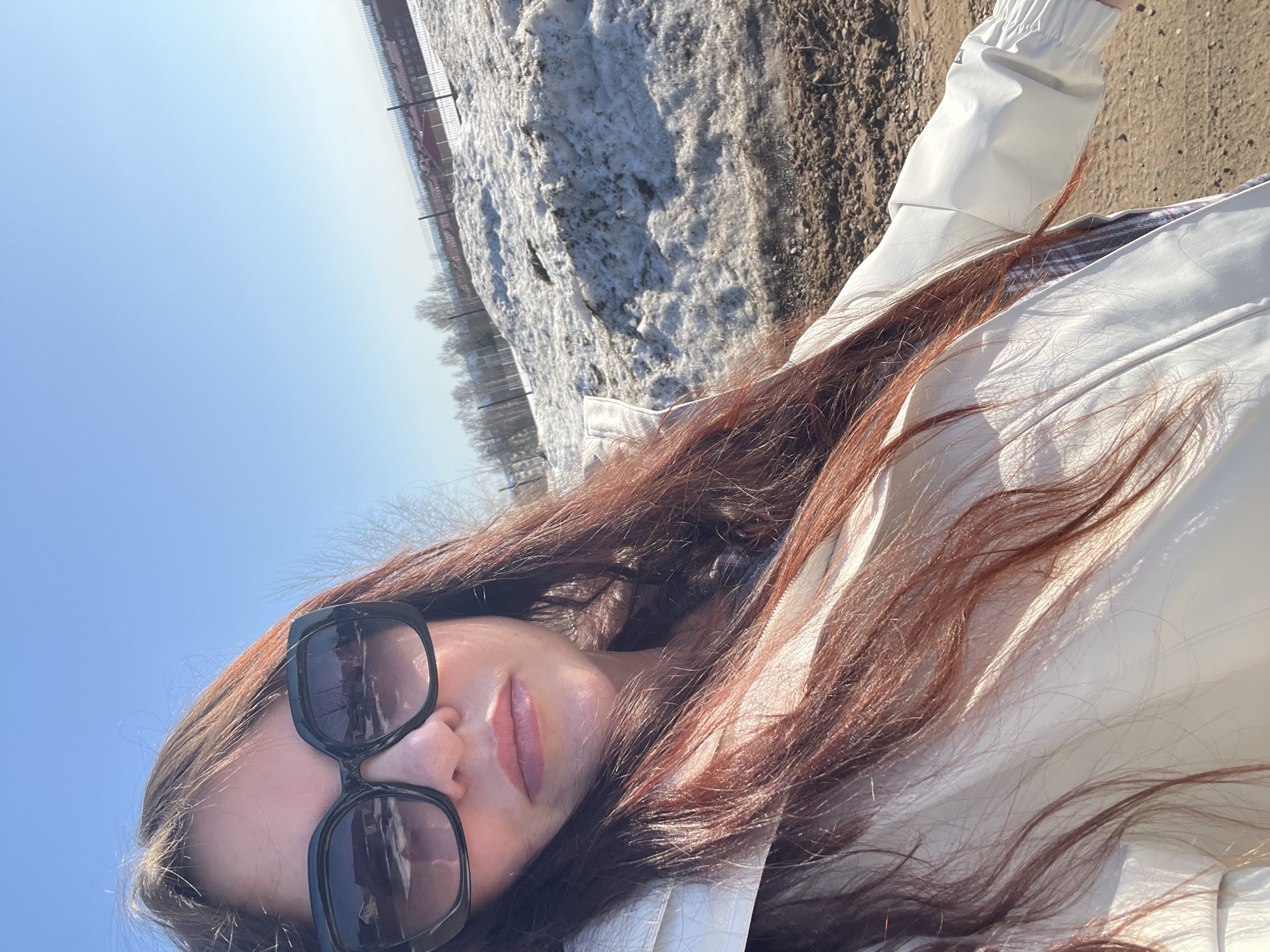 A woman with long, reddish-brown hair is wearing large, dark-rimmed sunglasses. She is wearing a light-colored jacket. The background shows a bright blue sky and a snow-covered area with trees and a fence in the distance. The scene appears to be outdoors during the daytime. The image does not contain enough information to verify the location in the city of Noviy, Russia.