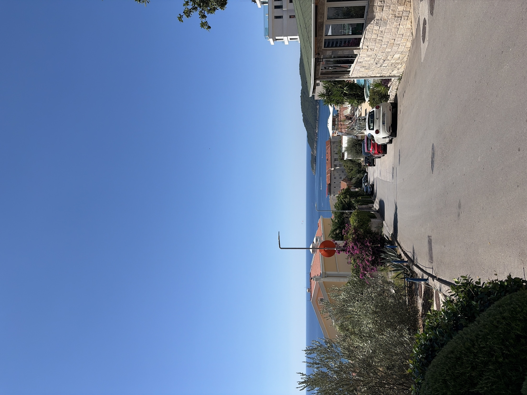 The image depicts a street in Bečići, Montenegro, with the Adriatic Sea visible in the background. The street slopes downwards towards the sea, lined with buildings and vegetation. On the left, there is a building with a yellow facade and an orange roof, accompanied by an olive tree and purple bougainvillea. Further down the street, several cars are parked, including a white Alfa Romeo. On the right, a stone building with a green roof and large windows is visible. The sky is clear and blue.