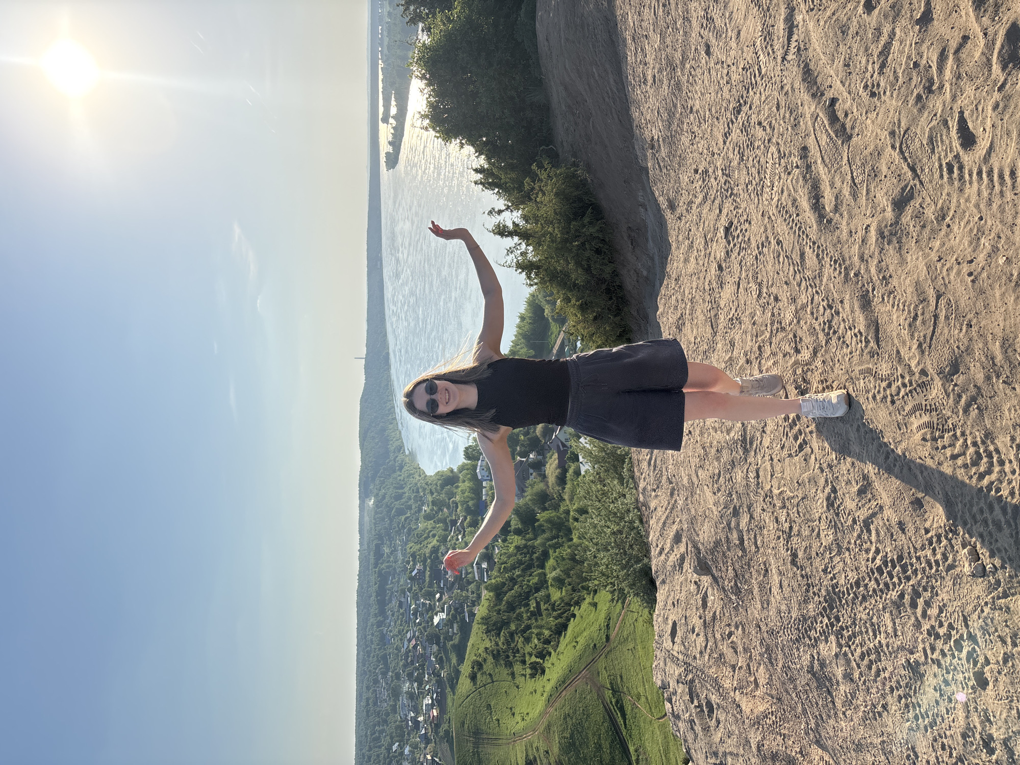 A medium shot, taken from a low angle, depicts a woman standing on a dirt ledge overlooking a vista. The woman, positioned in the center of the frame, wears a black tank top, dark shorts, sunglasses, and light-colored sneakers. Her arms are extended outwards.
The background features a river, green foliage, and buildings, consistent with the location being in Ольховка, Russia. The sky is bright blue, with the sun positioned in the upper left corner. The ledge the woman stands on appears to be made of sandy soil, marked with various footprints. The lighting suggests it is daytime.