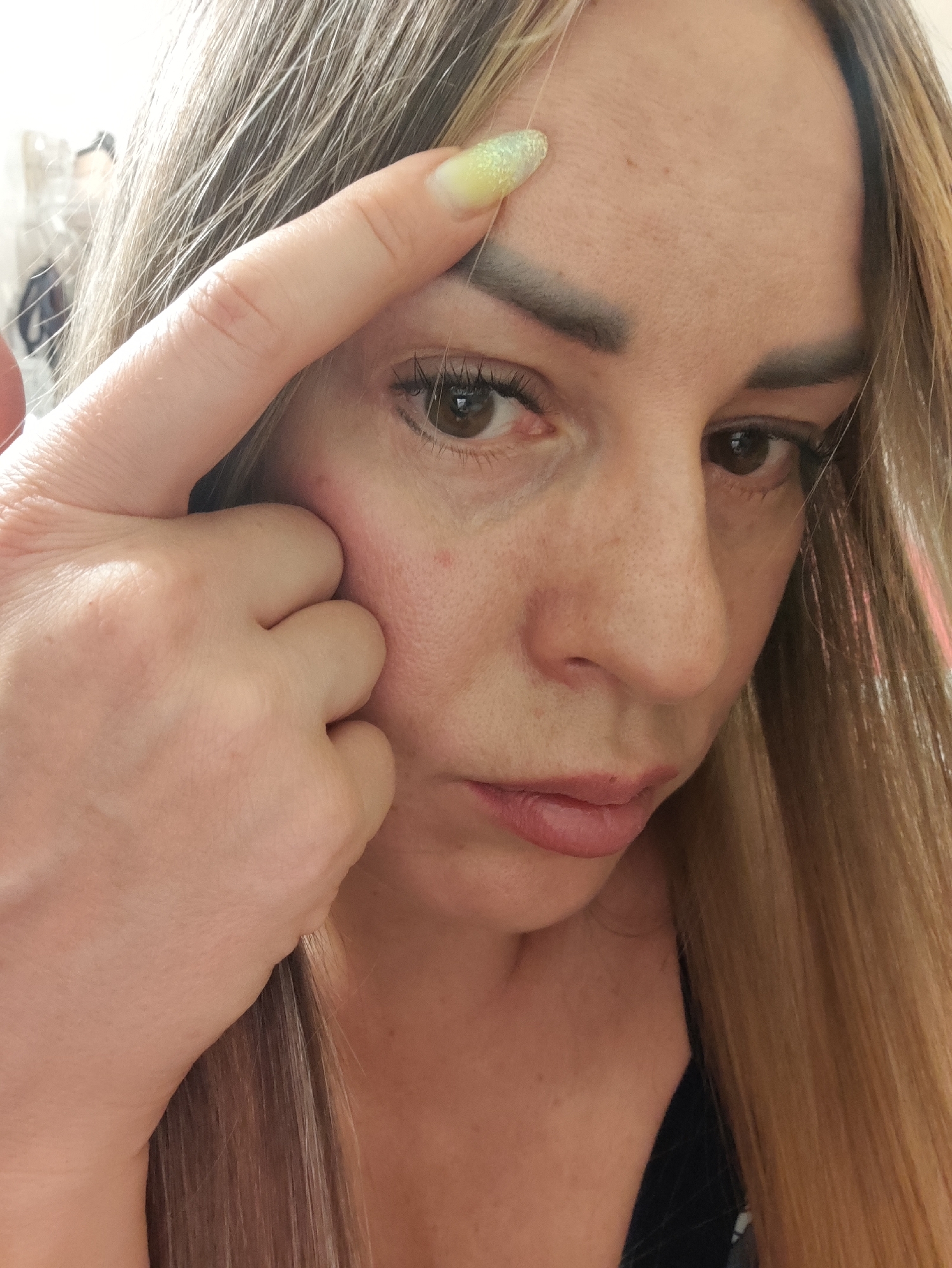 A close-up, eye-level, medium shot shows a Caucasian woman with long, blonde hair, her right hand extended to touch her forehead. Her left eye is partially obscured by her hand and hair, and her right eye is looking towards the camera. She is wearing light-colored makeup, including eyeliner and mascara. The background is out of focus and appears to be indoors, possibly a room with furniture and clothing visible. The overall lighting is soft and diffused. There is no identifiable information indicating the location in Almaty, Kazakhstan.