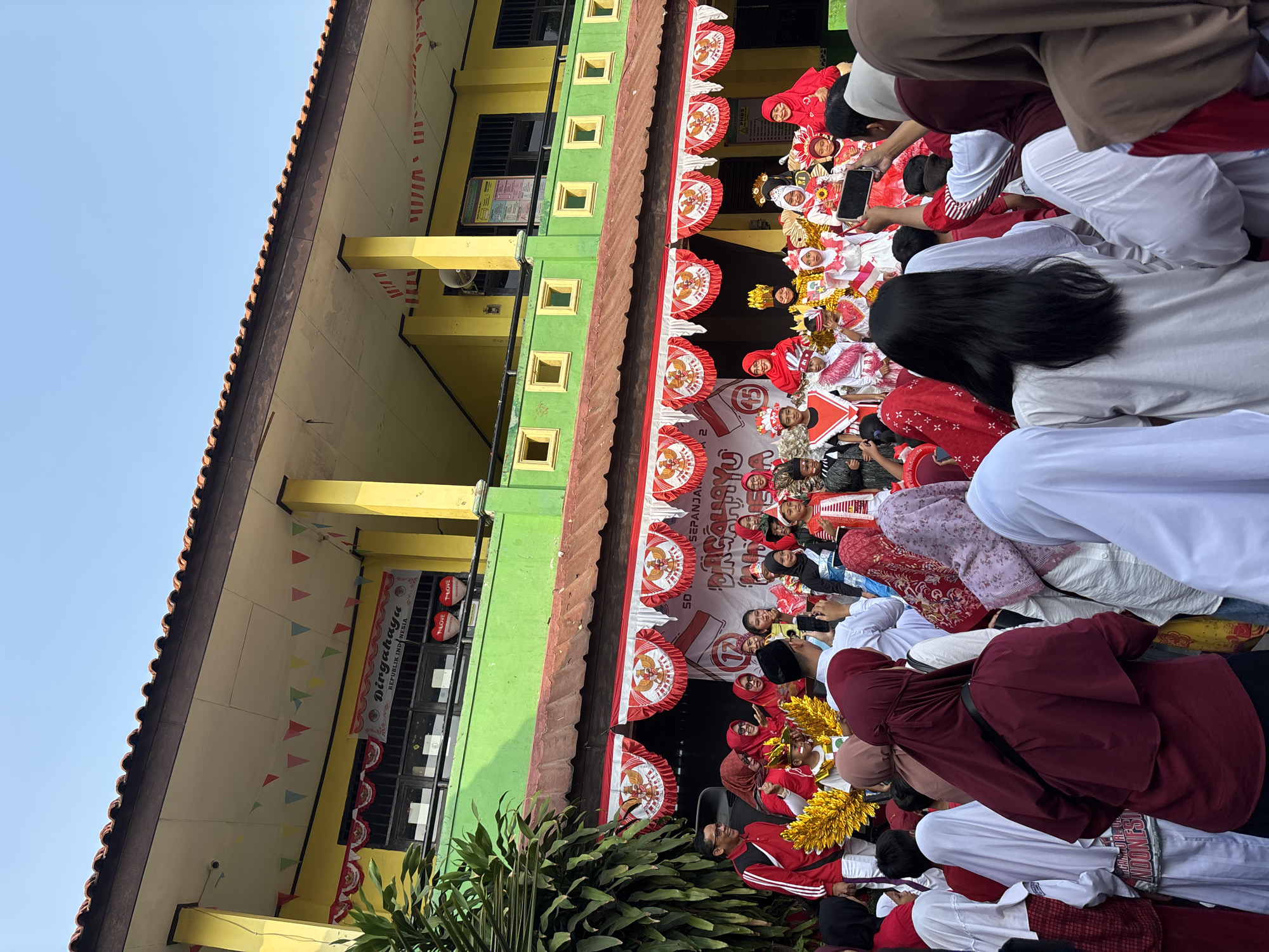 This is a medium-angle, full outdoor shot of an event taking place in front of a school building in Bekasi, Indonesia. The school building is painted yellow and green, with a tiled roof. Festive red and white banners are hung across the building and a stage area. A crowd of people, many wearing red and white or traditional Indonesian attire, are gathered to watch a performance. On the stage, children in elaborate costumes, some with gold wings and headdresses, are posing. The sky is clear and blue.