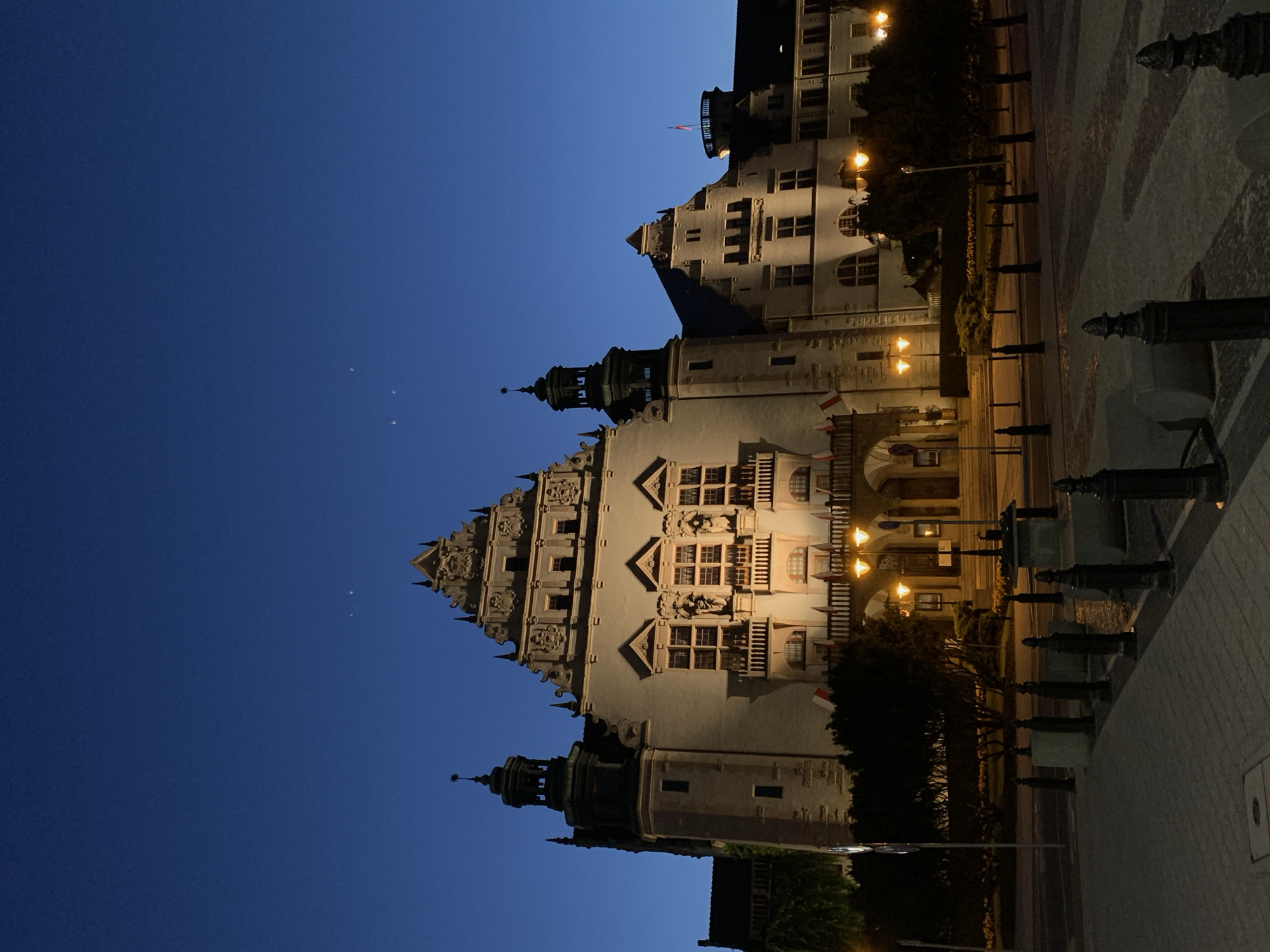 The image shows a nighttime view of the historic Collegium Minus building at Adam Mickiewicz University in Poznan, Poland. The sky is a deep twilight blue, with a few faint stars visible. The building itself is illuminated by warm artificial lights, highlighting its ornate architecture. The facade features multiple stories with arched windows, decorative carvings, and balconies. Two towers with green roofs flank the central section of the building.
In front of the building, there are trees and landscaping, also lit by streetlights. A paved plaza with bollards and some decorative stone elements is in the foreground. Several Polish flags are visible on the balconies of the building. The scene is quiet and still, suggesting a calm evening. There are no people visible in the image. The overall impression is one of historical grandeur and architectural beauty, presented in a serene nighttime setting.