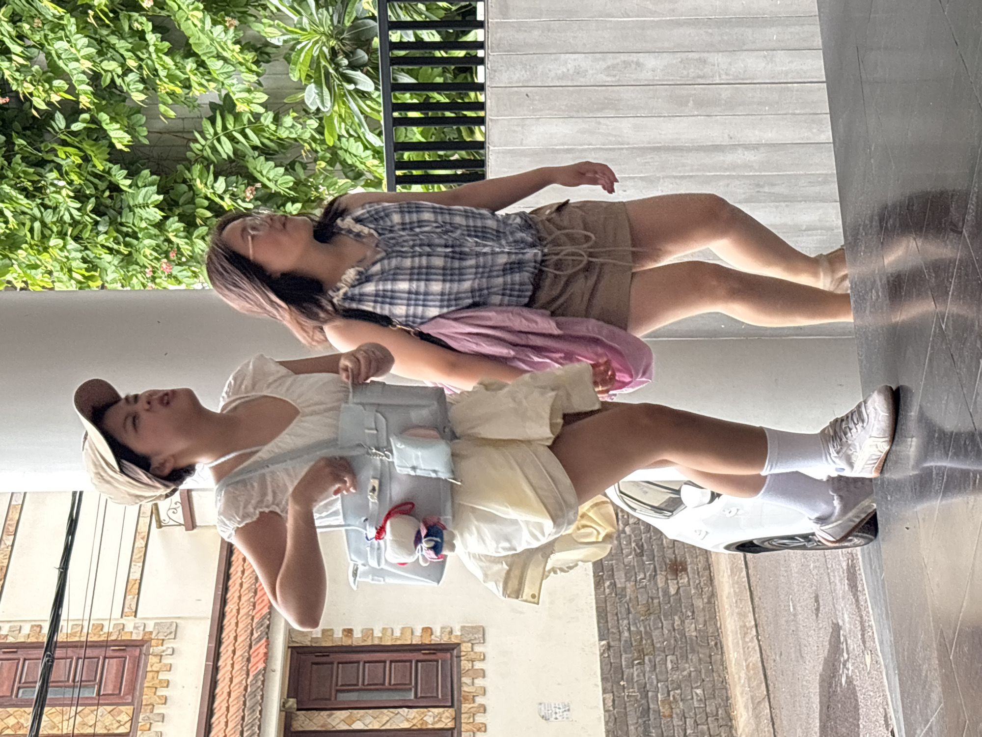 Two young women are walking outdoors. The woman on the left wears a white ruffled dress, a tan baseball cap, and a light blue handbag with a small white and red keychain. The woman on the right wears a blue and white checkered halter top, tan shorts, and carries a pink bag over her shoulder. They are walking on a tiled surface in front of a building with brick accents and a wall covered in green foliage. A white car is partially visible in the background. The scene appears to be a street in South Quy Nhon Ward, Vietnam.