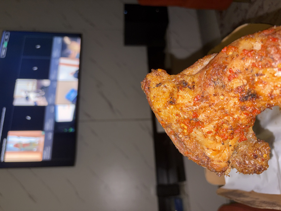 The scene depicts an indoor environment in the city of Igboefon, Nigeria. In the immediate foreground, a single, large, cooked chicken wing is prominently displayed, resting on white parchment paper within a light-brown cardboard takeout container. The chicken wing features golden-brown skin heavily coated with a vibrant orange-red spice mixture, exhibiting visible red chili flakes and dark, charred specks. The background reveals a light-colored wall, possibly white or off-white, with subtle gray veining, resembling marble or granite. Mounted on this wall is a flat-screen television displaying a grid layout consistent with a multi-participant video conference, though individual video feeds are blurred. Below the television, a dark, horizontal shelf or console is visible, along with a dark, vertical rectangular object to its right, likely an electronic device. A partial view of a reddish-orange object, consistent with a piece of furniture, is visible further to the right. The overall lighting suggests a dimly lit interior space.