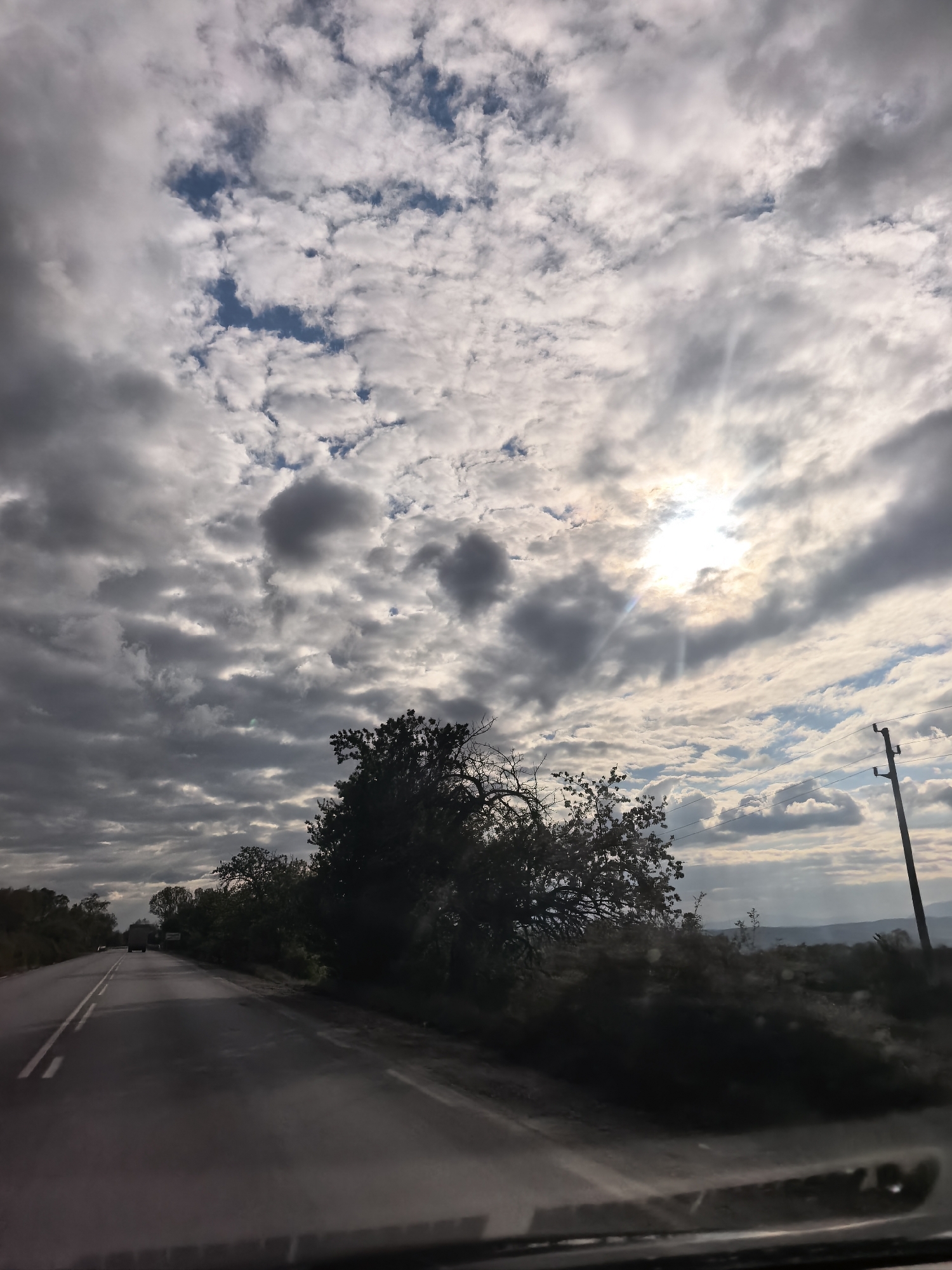 The image shows a view from inside a vehicle, looking out onto a rural road in Yahinovo, Bulgaria. The road has a double yellow line dividing lanes, and it is bordered by vegetation including trees and bushes on the right. A utility pole stands along the right side of the road. In the distance, the landscape appears somewhat hilly. The sky is predominantly covered with clouds, with patches of blue visible. The sun is obscured by the clouds, but its light is still visible through them.