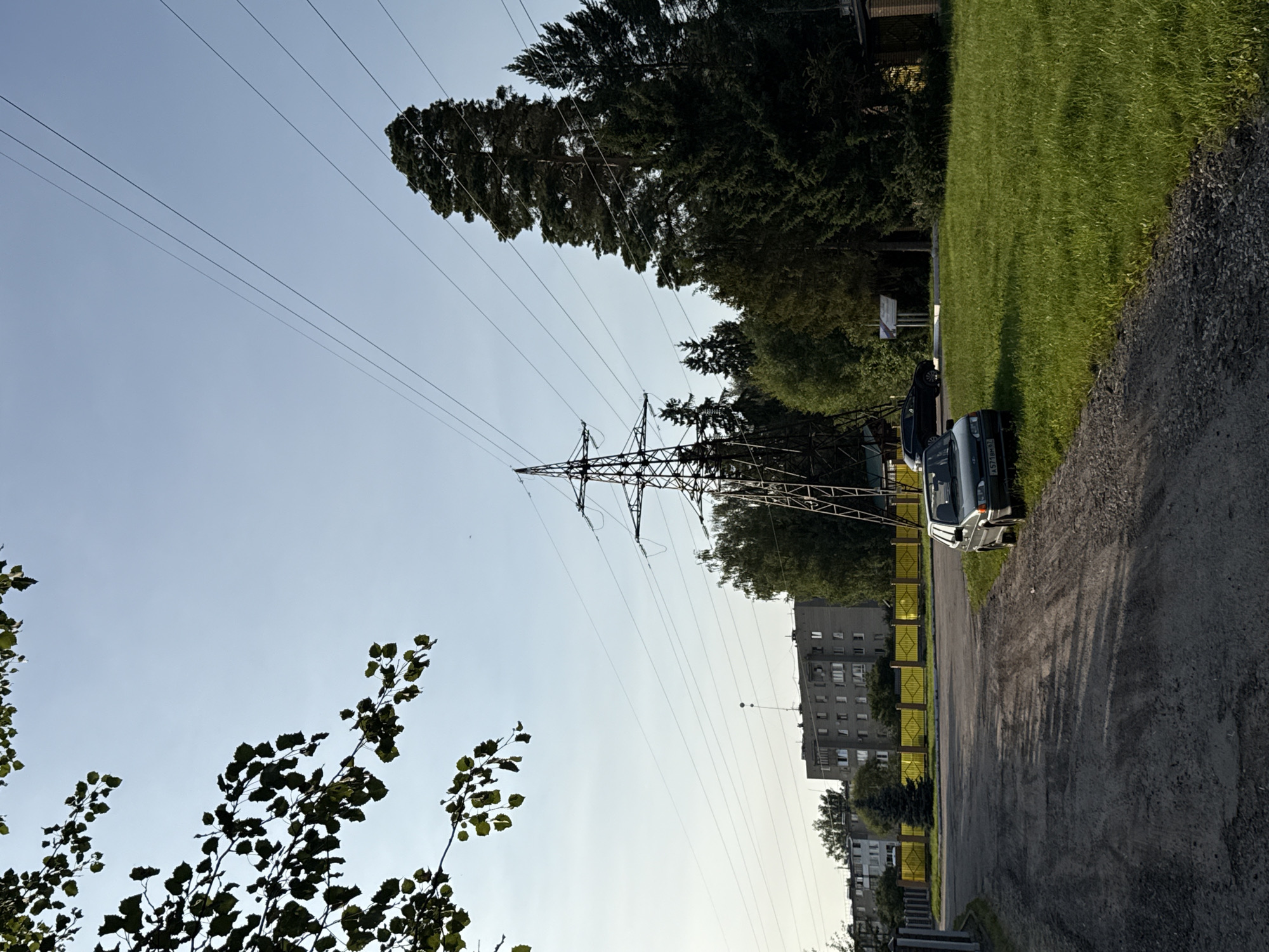 The image depicts a street scene in Volkovo, Russia, on a clear day. A dirt road curves from the lower left towards the center, with a dark sedan parked on the right side of the road. The car's license plate is visible. Beyond the road, a bright yellow fence runs horizontally, separating the road from a grassy area and buildings. In the background, several multi-story apartment buildings are visible, typical of Soviet-era architecture. A large electrical pylon stands prominently in the middle ground, with power lines stretching across the sky. Lush green trees, including tall pine trees, surround the pylon and extend towards the right side of the frame. The sky is a clear, pale blue.