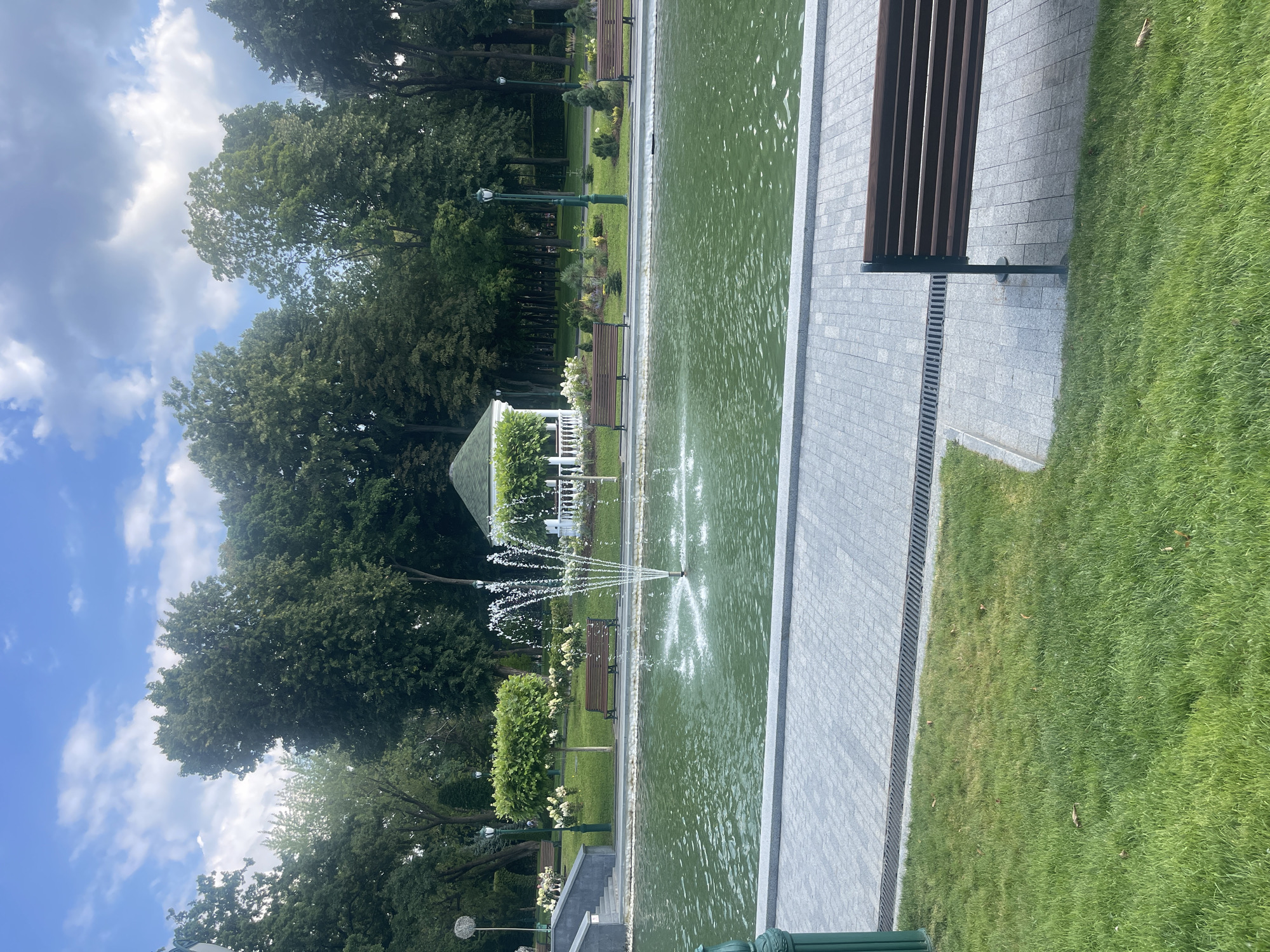 A full shot of a tranquil park scene on a cloudy day shows a vibrant green pond with a water fountain in the center, located in an urban park with lush trees and manicured lawns. The pond is surrounded by a grey stone walkway and a grassy embankment, with several wooden benches placed along the water's edge. In the background, a white gazebo with a green roof stands amidst decorative shrubs and trees, alongside lampposts and more benches. The sky is a mix of blue and white clouds, casting a soft light on the scene. There are no people visible in the image.