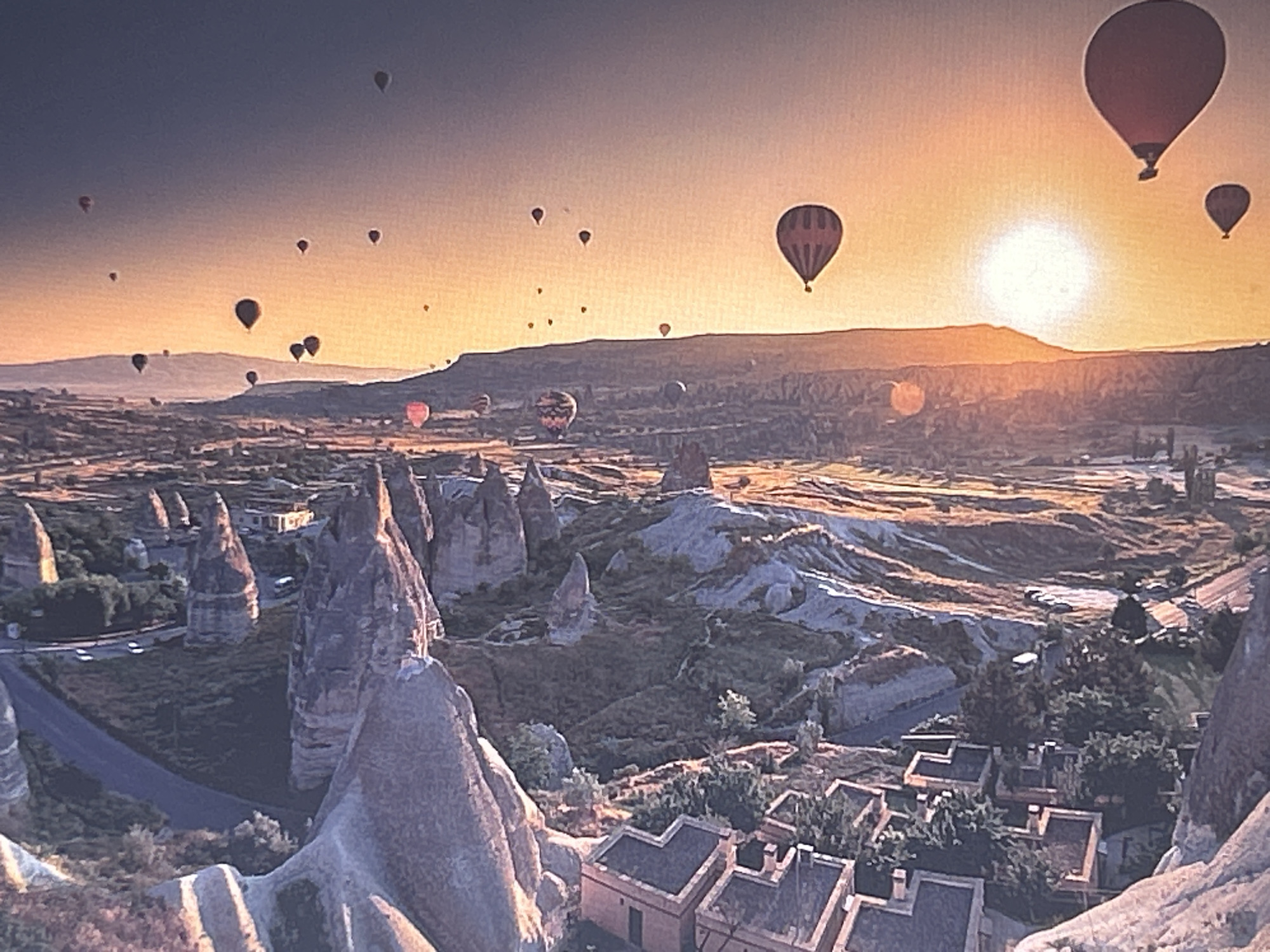This is a wide-angle, aerial shot of Cappadocia, Turkey, at sunrise, filled with hot air balloons. The sky transitions from a deep twilight blue at the top to a vibrant orange and yellow near the horizon, illuminated by the rising sun. Numerous hot air balloons of various sizes and colors are scattered across the sky, some appearing as silhouettes against the bright sun. The landscape below is characterized by unique geological formations, resembling fairy chimneys, covered in sparse vegetation and scattered with small buildings. A winding road is visible, along with some vehicles. The foreground shows a rocky, eroded terrain leading down to a valley with more rock formations and buildings. The scene captures a popular tourist activity in a surreal and breathtaking natural setting.