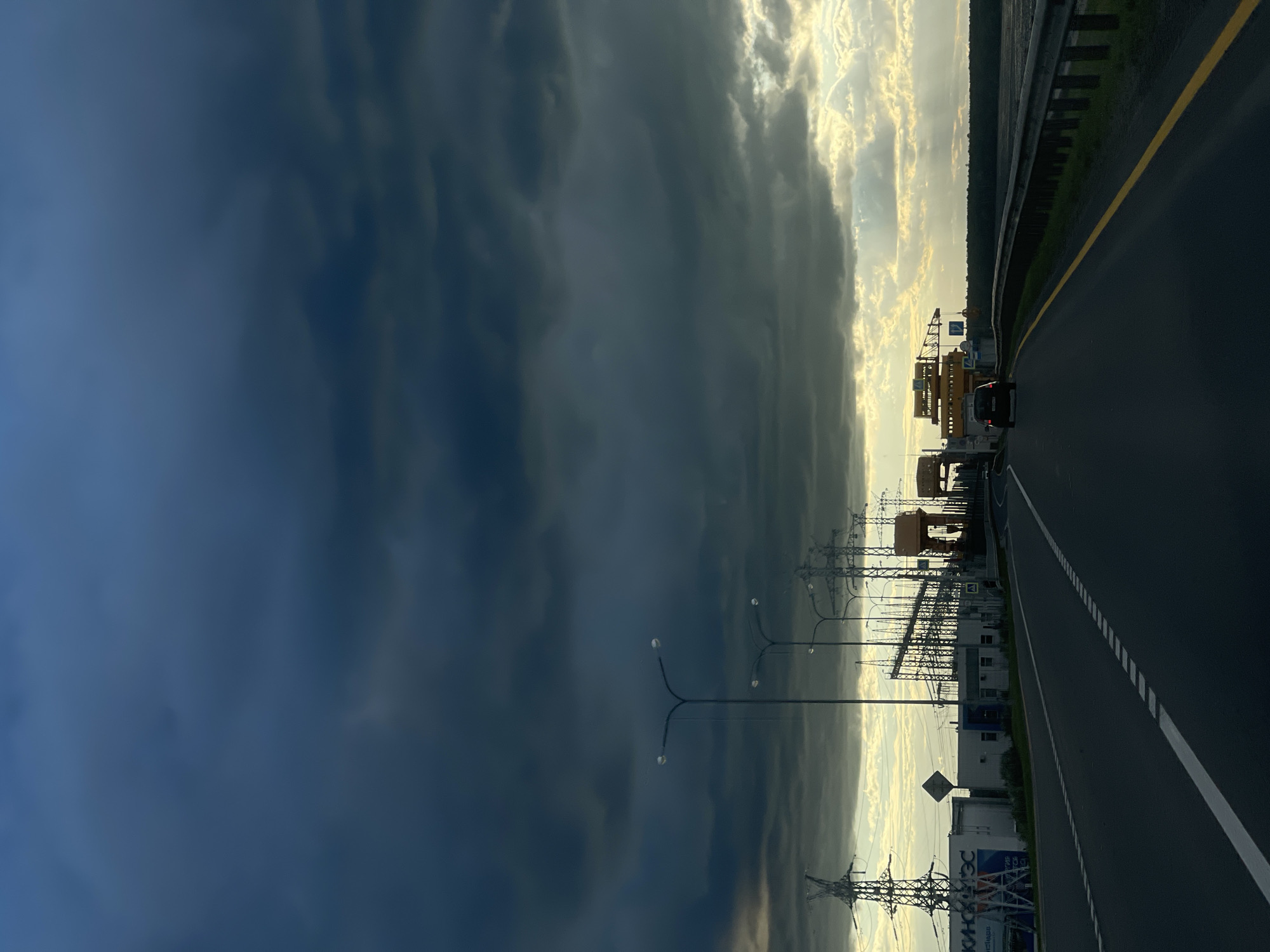 The image depicts a dramatic sky with dark, ominous storm clouds on the left and bright, illuminated clouds on the right, suggesting a transition between weather systems. A highway stretches into the distance on the right side of the frame, marked with white dashed lines and a yellow line on the shoulder. A black car is visible on the highway, appearing to be traveling away from the viewer.
Along the left side of the highway, a row of utility poles with multiple lights stands out against the dark sky. Further in the background, industrial structures, including what appear to be construction cranes and electrical substations with tall metal towers, are silhouetted against the brighter sky. The overall scene conveys a sense of movement and the immense power of nature, with the industrial elements contrasting with the natural dramatic sky.
The lighting suggests either early morning or late afternoon, with the sun's rays breaking through the clouds. The weather appears to be changing, with the dark clouds indicating an approaching storm, while the bright areas suggest clearing skies or the sun fighting through. There is a sign visible with some text on it, but it is not legible. Another sign in the background has Russian Cyrillic text that appears to read "РОСЭНЕРГОАТОМ" and another word below it starting with "ЭНЕРГ" which is likely related to energy.