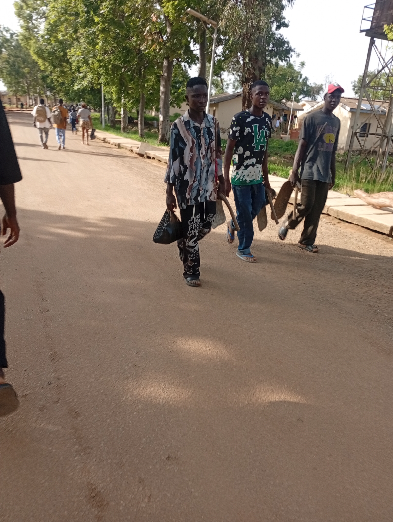 The image depicts three young men walking on a dirt road in Rafin Par, Nigeria. The man on the left wears a black t-shirt and dark pants. The middle man is dressed in a patterned shirt and dark pants, carrying a black plastic bag in his left hand. The man on the right wears a dark t-shirt, dark pants, and a red baseball cap. He is carrying a shovel in his right hand and a curved wooden tool in his left. The road is lined with trees and scattered buildings in the background. The overall scene is a casual outdoor setting.