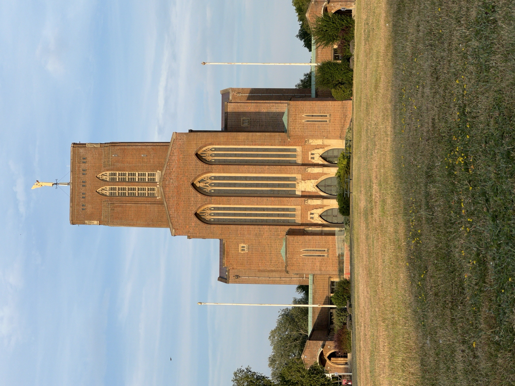 This is a vertically oriented photograph of Guildford Cathedral in Guildford, United Kingdom. The cathedral is a large brick building with Gothic-style arched windows. A flagpole stands to the left of the cathedral, and a yellow structure resembling a weather vane or finial is visible at the top of the building. The sky is a clear blue with some wispy clouds. The foreground is a grassy field with some small yellow flowers. Trees and bushes are visible behind the cathedral. The lighting suggests it is either early morning or late afternoon, casting long shadows. The scene is peaceful and serene. No people are visible in the image. There is a red sign visible at the bottom right corner of the image.