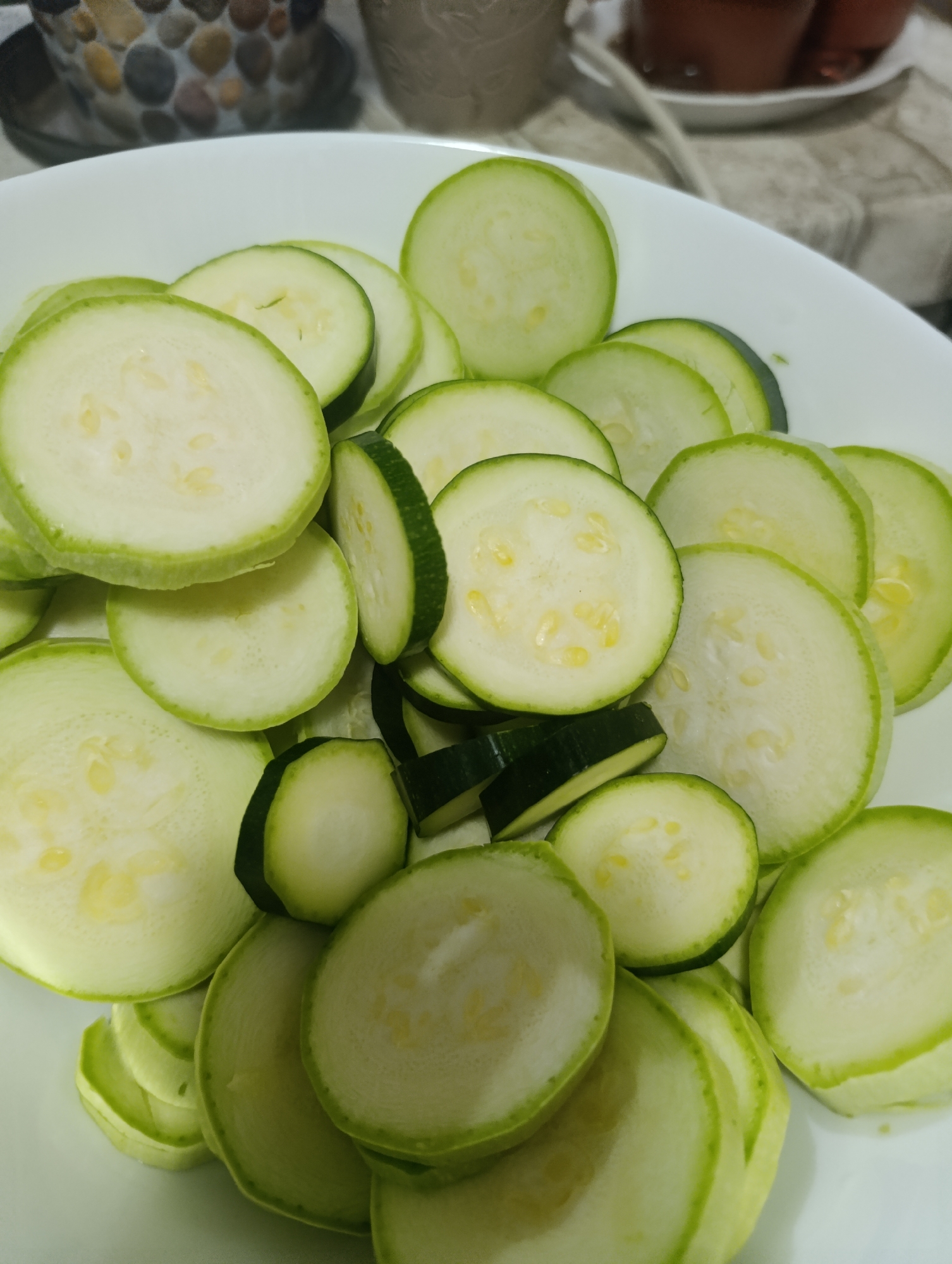 A white ceramic bowl is filled with sliced zucchini. The zucchini slices are round and thin, with a light green skin and white flesh speckled with small yellow seeds. Some slices are whole, while others are partial. The bowl is positioned on a light-colored surface, likely a kitchen counter. In the background, out of focus, are blurred objects including what appears to be a decorative ceramic pot with stones and a dark liquid in a cup or bowl. The lighting suggests an indoor setting. The image is a close-up of the zucchini, filling most of the frame. There is no indication of the city of Kyustendil, Bulgaria, in the provided image.