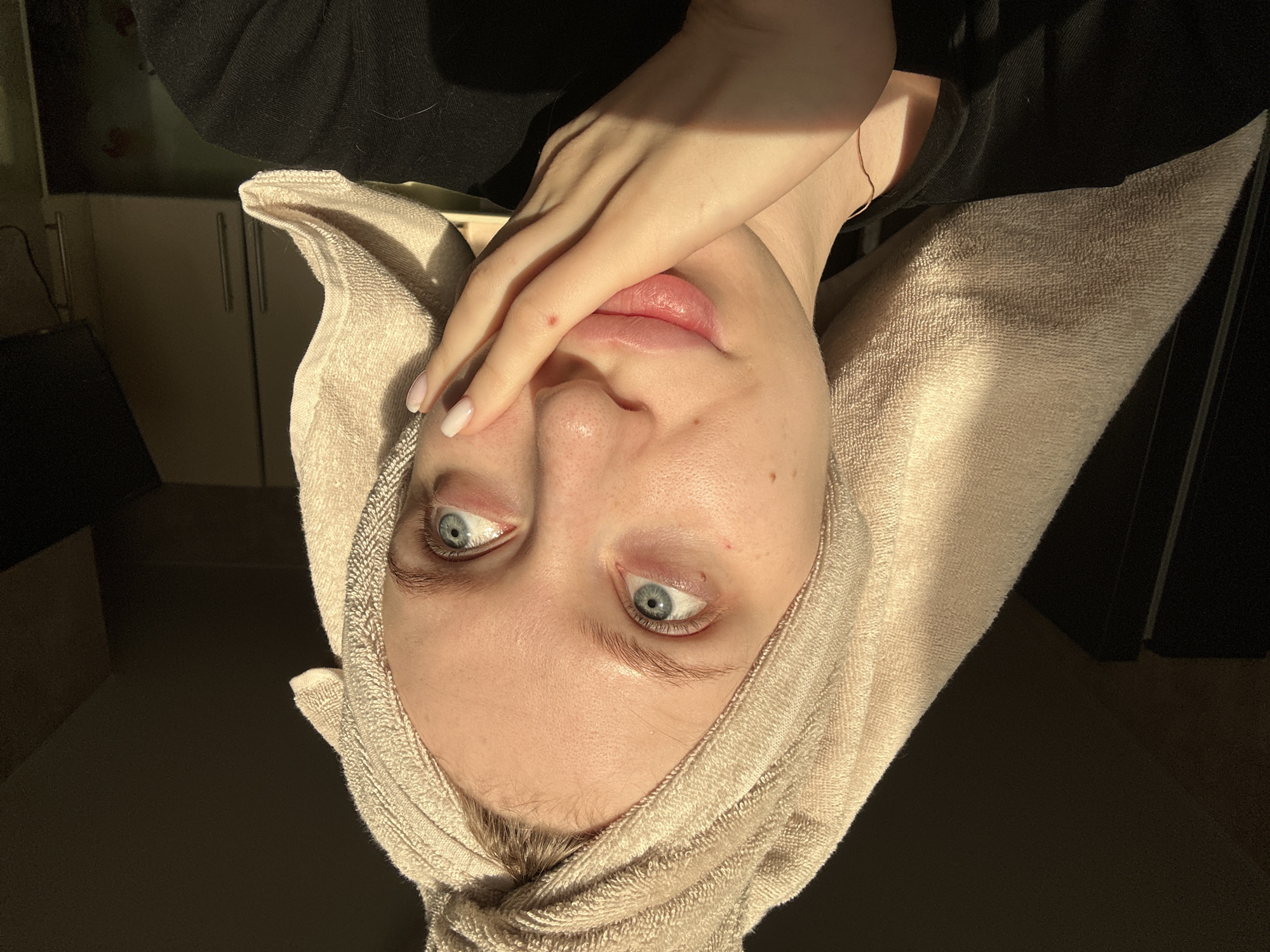 A close-up, inverted portrait of a woman with fair skin and striking blue eyes, possibly taken indoors, with a background that appears dark. She is wearing a beige towel wrapped around her head, and her right hand is raised, partially covering her face. Her nails are painted white. The lighting appears to be coming from the lower right, casting shadows on the towel and her face. There is no discernible indication of a specific location, especially the city of Chaikovsky, Russia.