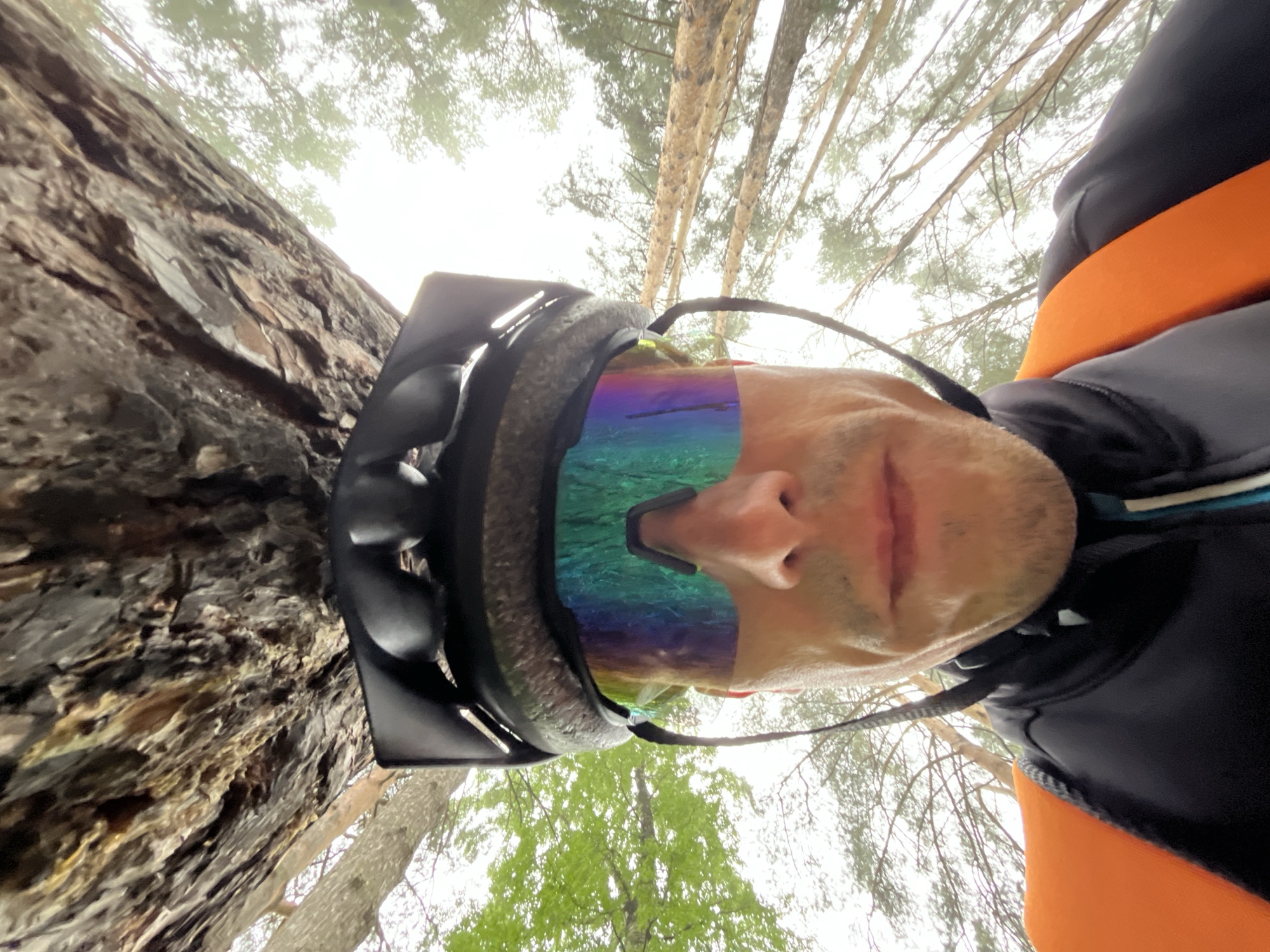 An inverted, wide-angle shot captures a man in outdoor gear within a forest environment, with the perspective looking upwards. The central subject is a man wearing a dark, robust helmet and large, iridescent sports goggles that reflect a vibrant spectrum of blues, greens, and purples. His face, visible from the nose downwards, shows light stubble, and his expression appears neutral. He is dressed in a dark, long-sleeved top accented with a bright orange band or stripe across the chest and shoulder.
On the left side of the frame, a massive tree trunk with rough, deeply textured bark is prominently featured, consistent with a pine or similar coniferous tree. The background reveals a dense canopy of tall, slender tree trunks and branches silhouetted against a bright, overcast sky, indicating it is daytime. Sparse green foliage is visible among the branches, suggesting a natural, wooded setting. The unusual, low-angle perspective, combined with the helmet and goggles, suggests the man is engaged in an outdoor activity such as cycling or mountain biking, and the image is likely a selfie taken with a wide-angle lens. No discernible text is visible in the image.
