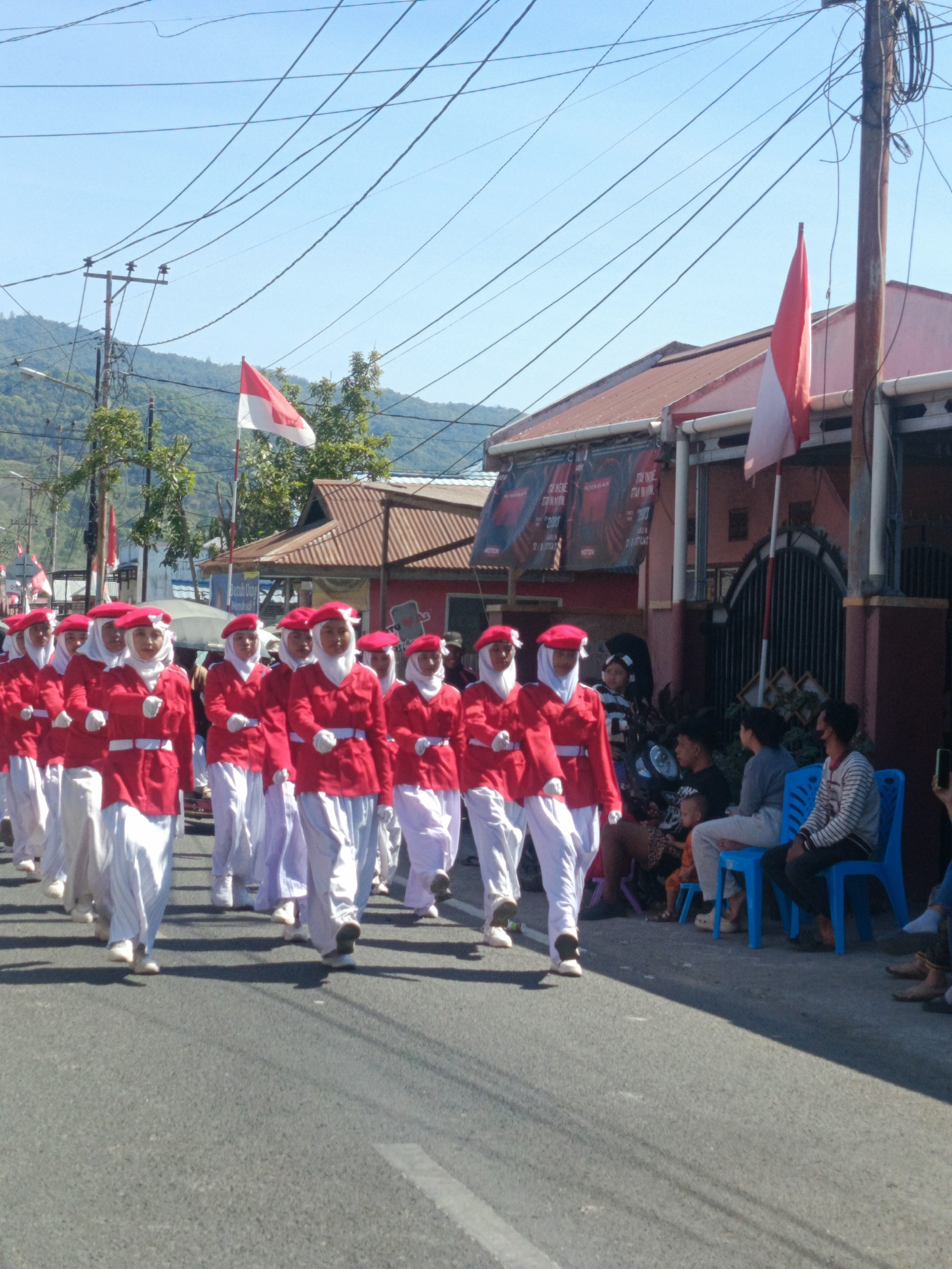 A procession of young people, likely students, wearing matching red jackets and white pants, walk in formation down a paved street. They are also wearing red berets and white hijabs. The day is sunny, with a clear blue sky overhead. Buildings line the street, and trees are visible in the background, with a hillside in the distance. Indonesian flags, red and white, are visible on poles and held by individuals in the procession. Spectators are seated on chairs along the side of the road, observing the event. The scene appears to be a parade or a formal march.