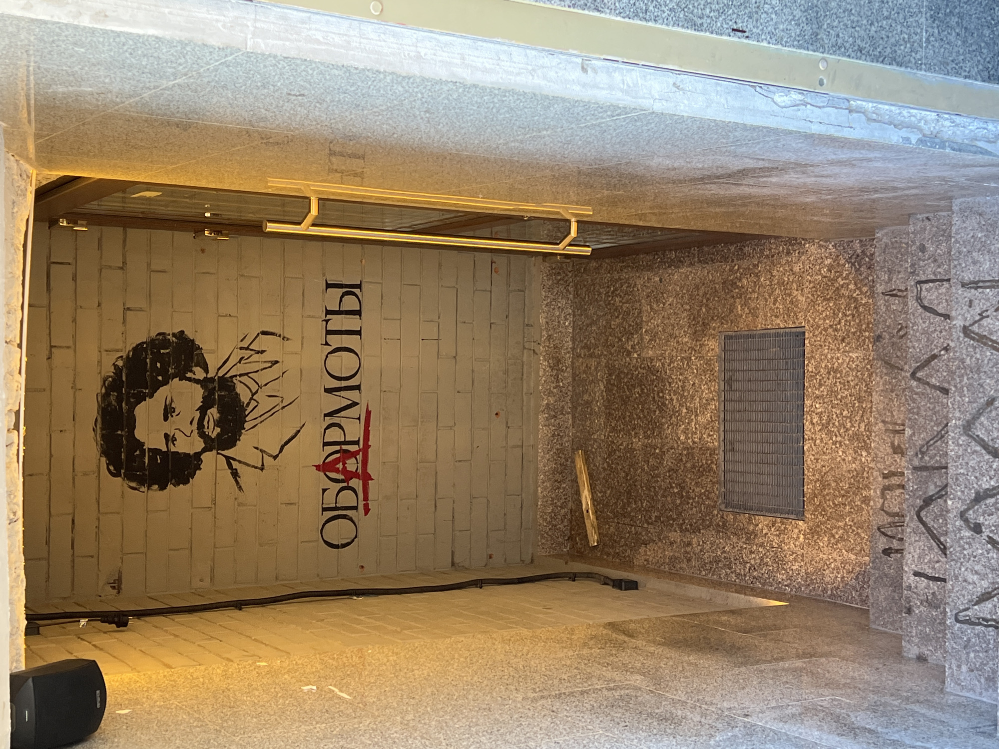 The image depicts an underpass or entrance to a building in Saint Petersburg, Russia. On the left wall is graffiti, including a stylized portrait with a beard and the Cyrillic word "ОБАРМОТЫ" written vertically, with a red line crossing it out. A black speaker sits on the floor. Overhead is a suspended light fixture with a rectangular shape. The right wall is a granite wall with a ventilation grille. The granite stairs are on the right side with graffiti. The floor appears to be tiled.