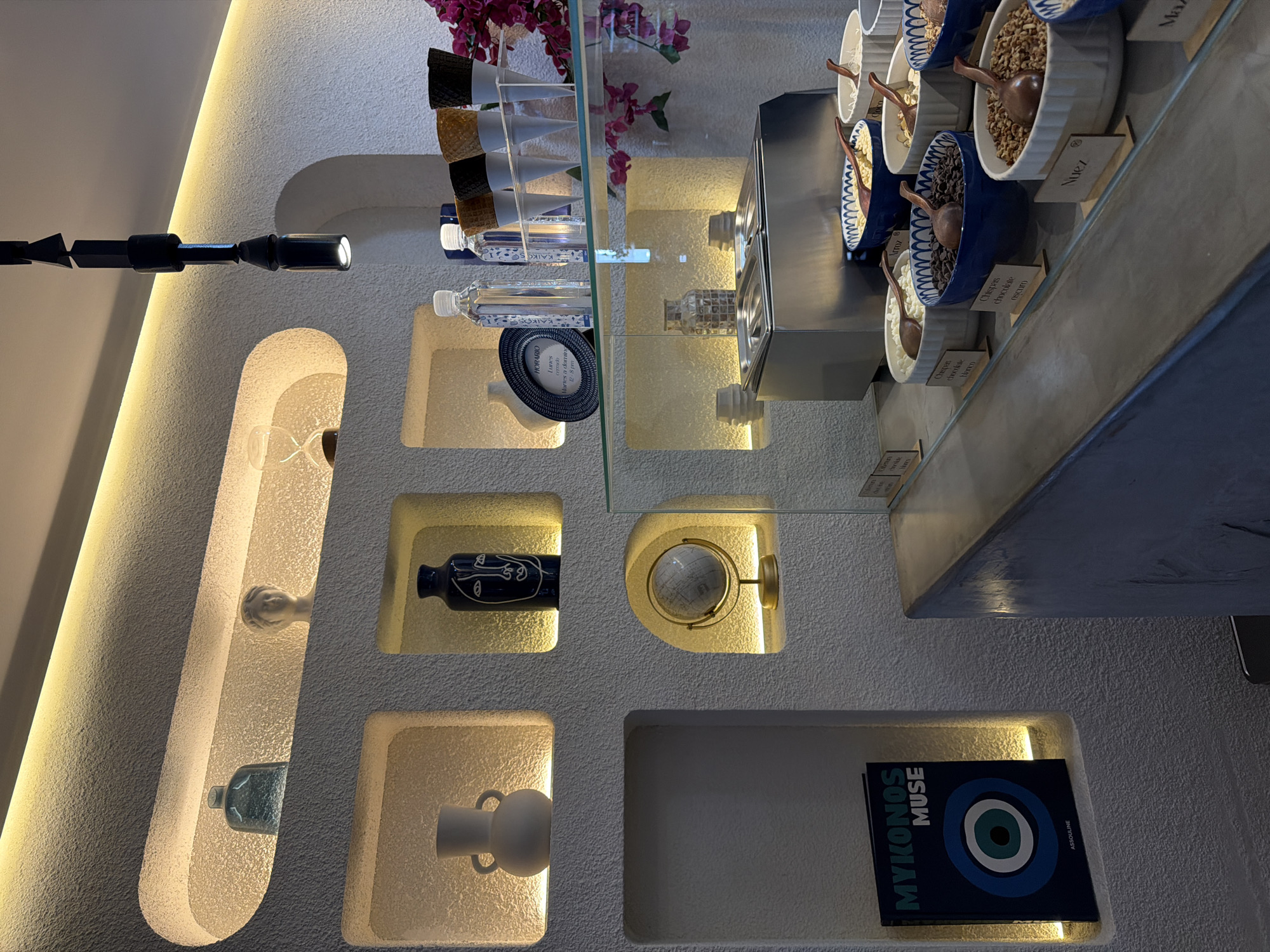 An interior shot depicts a modern and aesthetically designed commercial space, likely a dessert shop or cafe, with a minimalist white textured wall adorned with various illuminated display niches. The image appears to be rotated 90 degrees clockwise, showing the main counter on the right side.
The left side of the wall features a long, black, segmented light fixture. Below it, several recessed niches are illuminated with warm light. These niches hold decorative items, including an hourglass-shaped object, a small bottle of liquid, a dark blue vase with an abstract white face design, a small white handled vase, and a small golden globe on a stand. Towards the bottom right, a rectangular niche contains a book titled "MYKONOS MUSE" by Assouline, featuring a prominent blue and white "evil eye" symbol.
On the main display counter, several empty ice cream cones, including waffle and black varieties, are arranged in clear holders. Two clear water bottles, one labeled "KAIKOS," are also present. A blue and white ceramic dish, with text visible, is on the counter. The text reads "HORARIO" (schedule), "Lunes cerrado" (Monday closed), and "Martes a dom" (Tuesday to Sunday).
Below the glass counter, a silver-toned serving station is surrounded by several white and blue ceramic bowls filled with various toppings, each with a small wooden scoop. Wooden labels identify the toppings: "Chispas chocolate Blanco" (white chocolate chips), "Arroz" (rice), "Chispas chocolate oscuro" (dark chocolate chips), and "Nuez" (nut). Another partially visible label hints at "Ma..." (possibly marshmallows or corn). A bouquet of pink flowers adds a touch of color in the background. The setup strongly suggests an ice cream or frozen yogurt bar where customers select toppings. The overall ambiance is clean, inviting, and subtly themed, possibly with a Mediterranean influence given the "Mykonos" book and evil eye symbol.