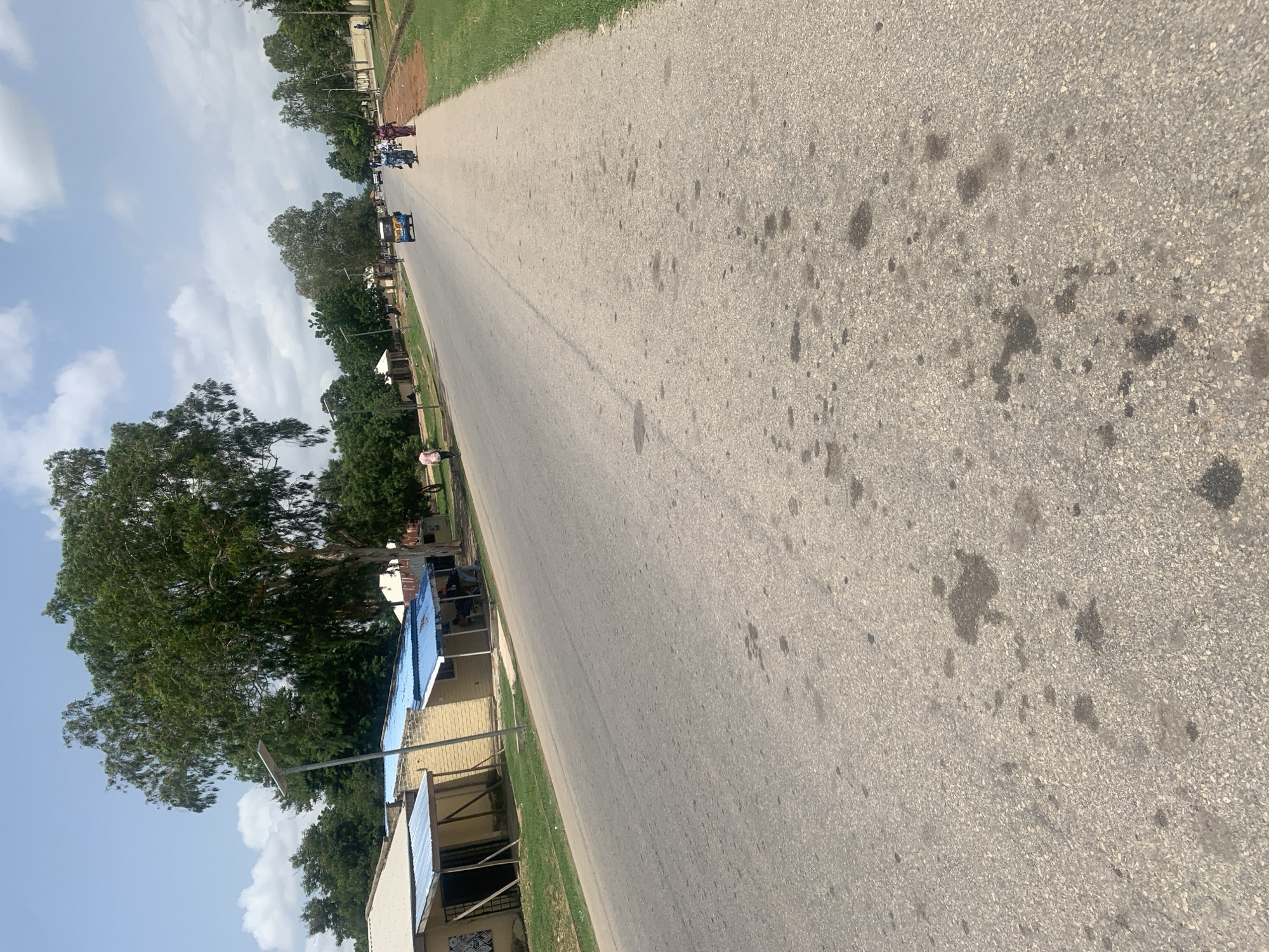 The image depicts a street scene in Jalingo, Nigeria, viewed from a low angle and rotated. The foreground is dominated by a grey asphalt road with dark oil stains. To the left, a grassy verge lines the road, with a row of buildings behind it. These buildings have yellow brick walls and corrugated metal roofs, some of which are blue. Trees, including a large one with dense foliage, are interspersed among the buildings. Further down the road, several people are visible, some standing and others riding in a blue and yellow motorized tricycle, commonly known as a "Keke Napep." The sky is partly cloudy, suggesting it is daytime, likely mid-morning or afternoon, given the bright light. The scene conveys a sense of everyday life and activity on a street in Nigeria.