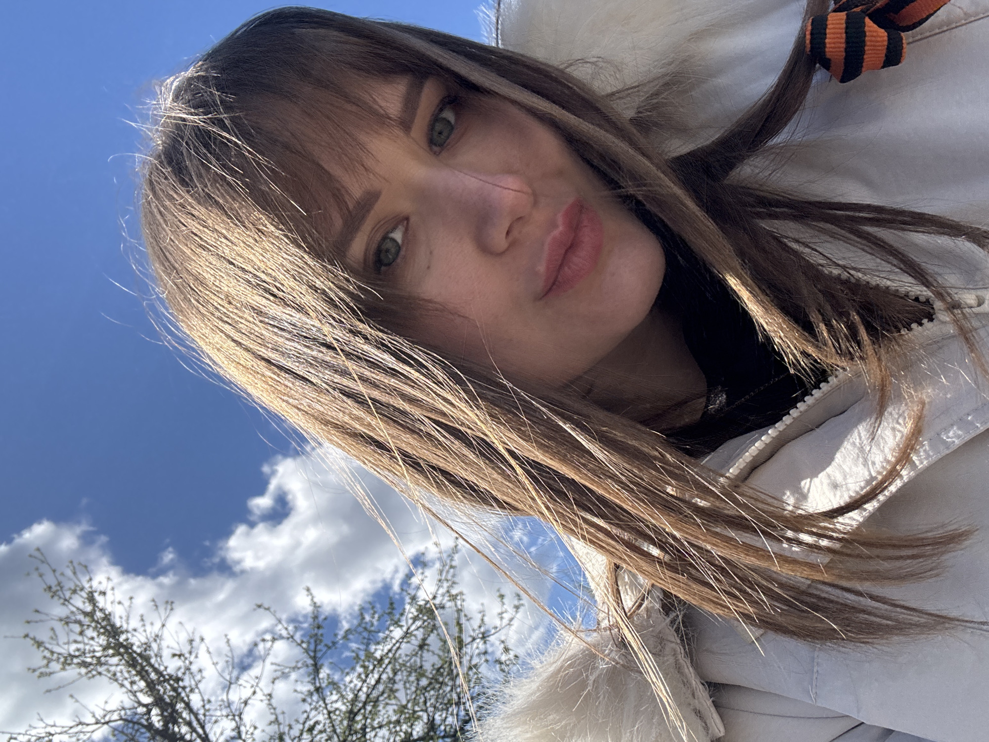 A young woman with brown hair and bangs takes a selfie against a background of a blue sky with white clouds and tree branches. She is wearing a white jacket with fur trim around the hood. An orange and black striped ribbon is attached to her jacket. The photograph was taken outdoors in Chaikovsky, Russia.