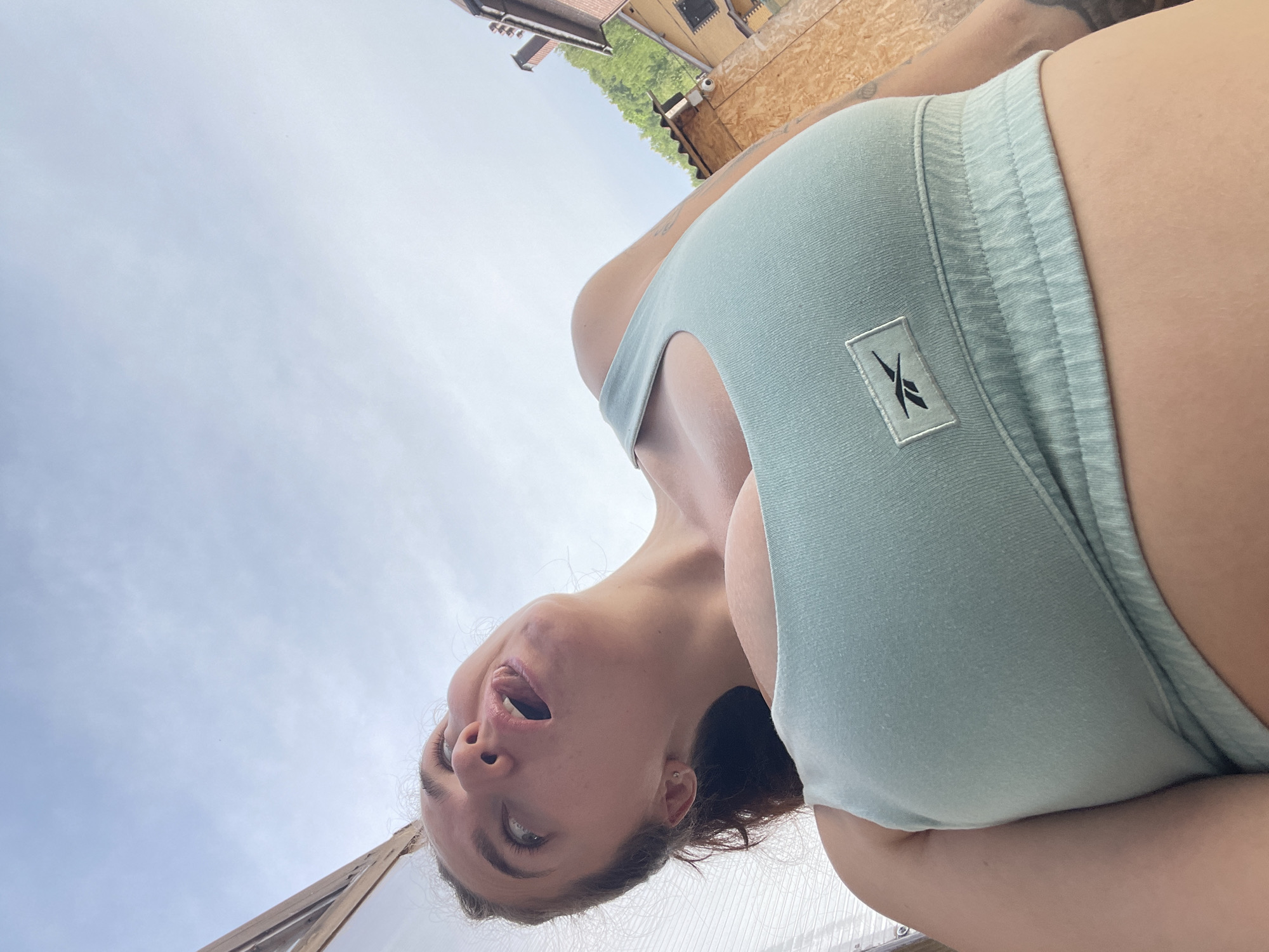 A woman is facing the camera with her head tilted back, mouth open. She is wearing a light blue athletic top with a Reebok logo and matching shorts. Behind her is a sky with light clouds and a building with wood paneling and what appears to be a security camera. Some kind of roof or covering is visible at the bottom of the image. The location is unknown.