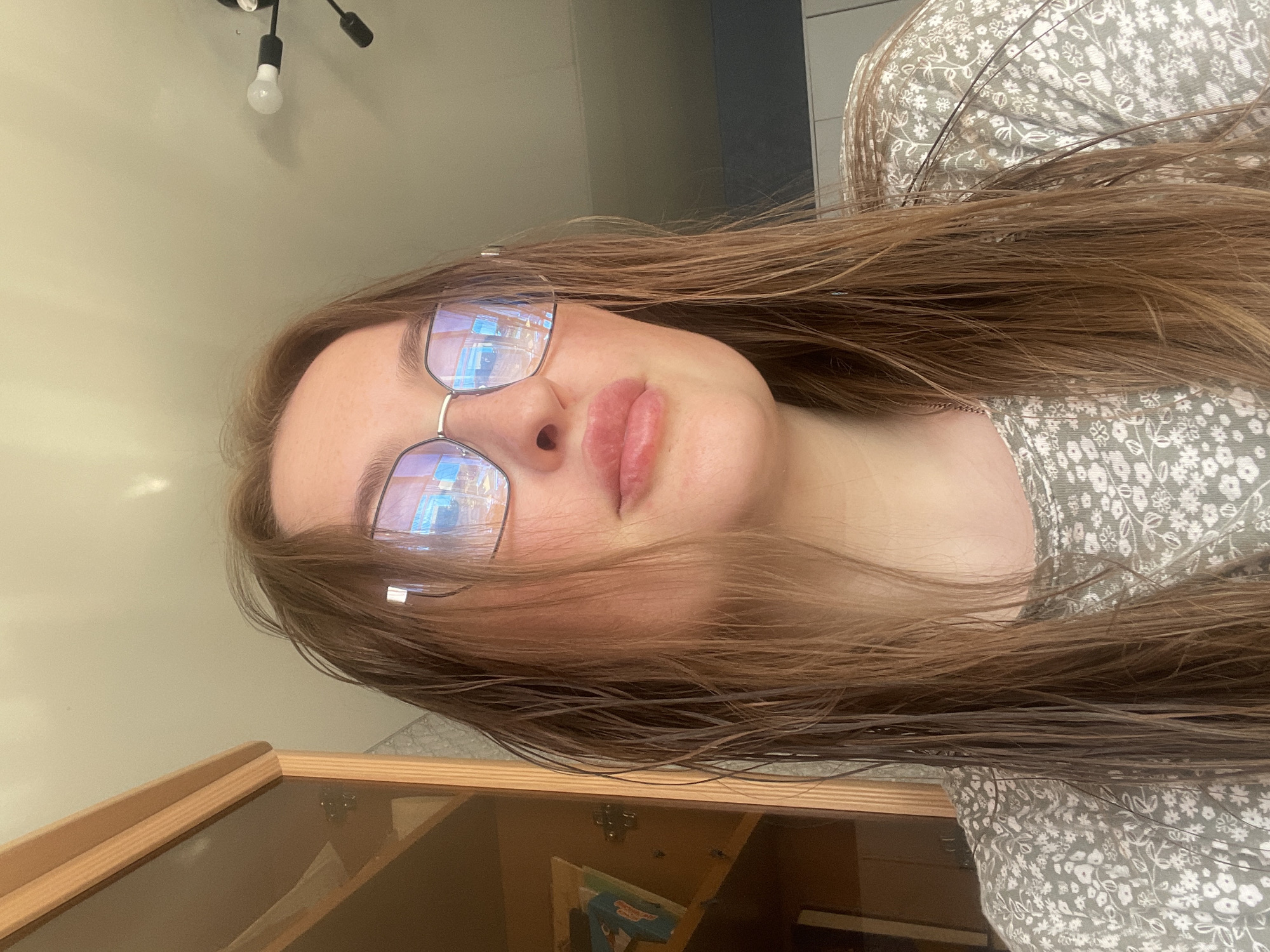 The image shows a young woman indoors, close-up, looking upwards towards the camera. She has long, wet, light brown hair, and is wearing glasses with a light-colored frame. Her lips are slightly parted. She is wearing a light green, floral-patterned shirt. The background includes part of a wooden cabinet to the left and a light-colored ceiling with a light fixture. There is no identifiable information in the image to suggest a specific location in the city of Сырково, Russia.