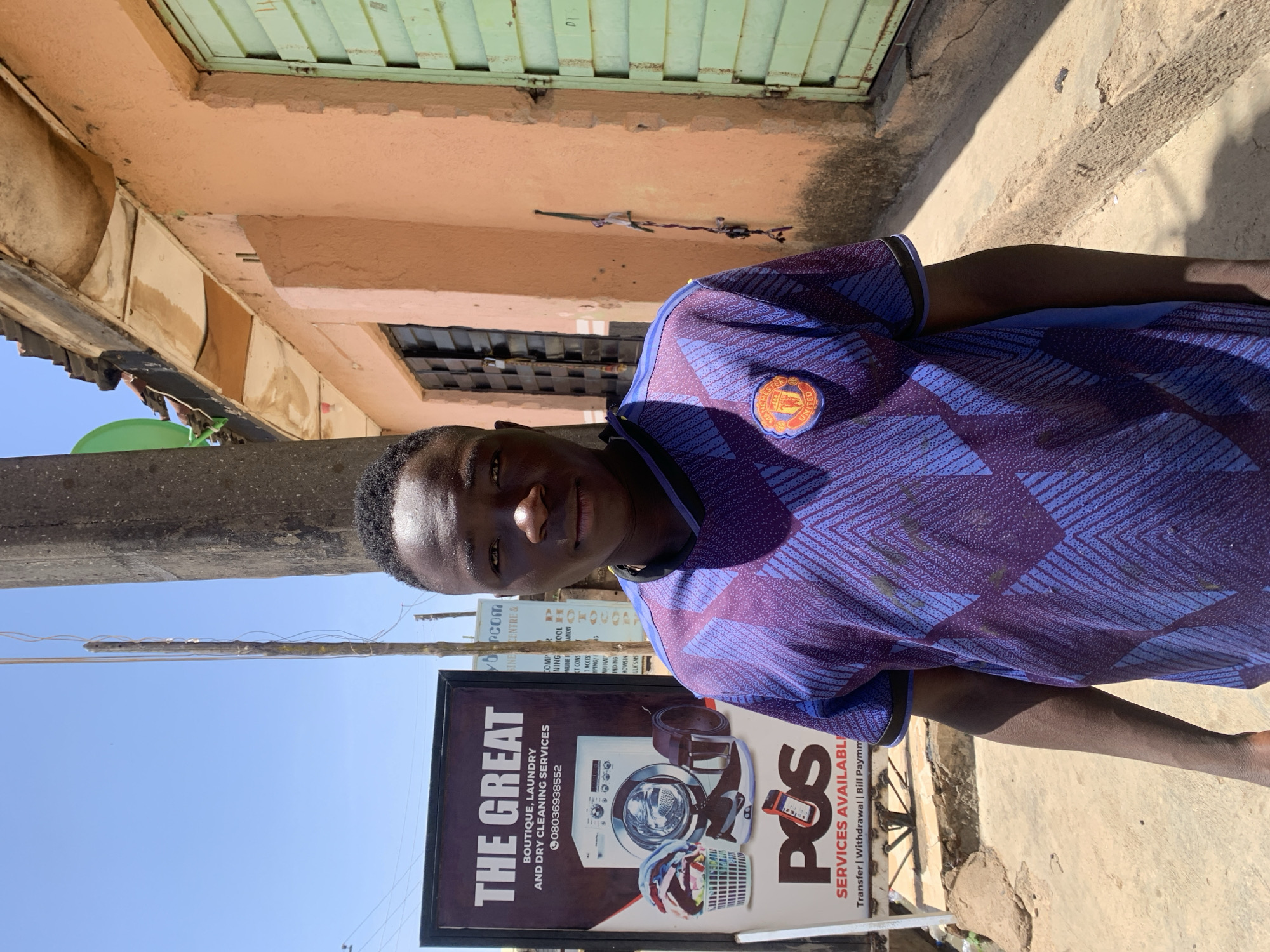 A young man stands in front of a business advertising laundry and POS services. The signage reads "THE GREAT BOUTIQUE, LAUNDRY AND DRY CLEANING SERVICES". The location is likely a commercial area in Jos, Nigeria, based on the text of the sign, the visual style of the business, and the physical appearance of the individual. The man is wearing a blue and purple Manchester United jersey and is facing the camera. Behind him is a peach-colored building with a weathered green door and other buildings in the background. A street lamp is visible. The overall environment appears to be an outdoor street setting.