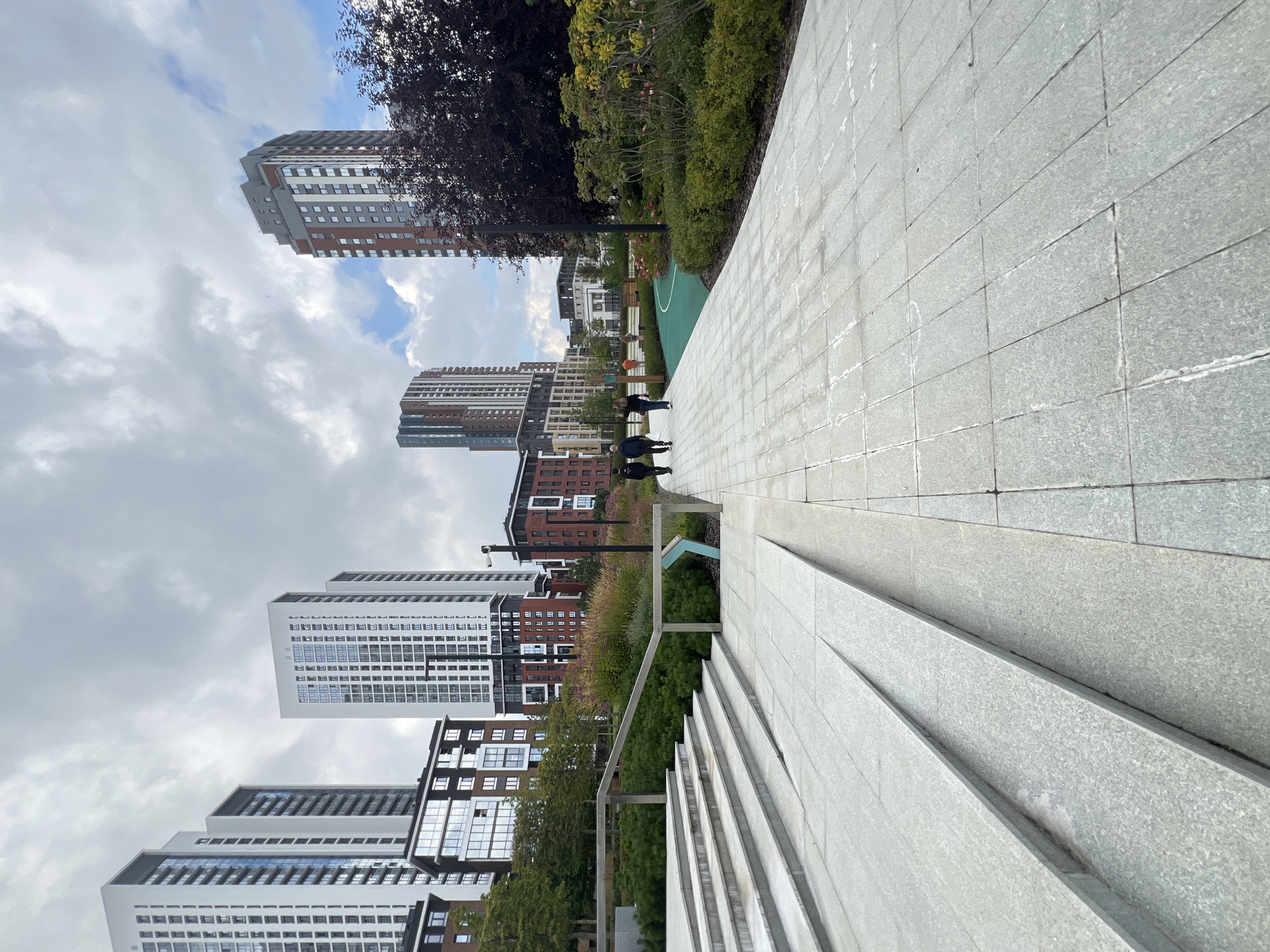 In Yekaterinburg, Russia, a wide paved walkway curves gently to the right, leading toward a cluster of modern high-rise buildings under a cloudy sky. To the left, a wide set of light-colored granite stairs descends, flanked by a metal handrail. Beyond the stairs, a dense green shrubbery area is visible. The walkway is made of light grey square tiles. Several people are walking away from the camera on the path; three individuals are in the foreground, and two more are further ahead. In the background, the buildings are a mix of white, red brick, and dark grey facades, with many windows. To the right of the walkway, lush green landscaping with some flowering bushes and a dark-leaved tree create a park-like setting. A distinct green track or pathway is visible in the mid-ground, curving into the landscape. The overall scene depicts a well-maintained urban outdoor space.