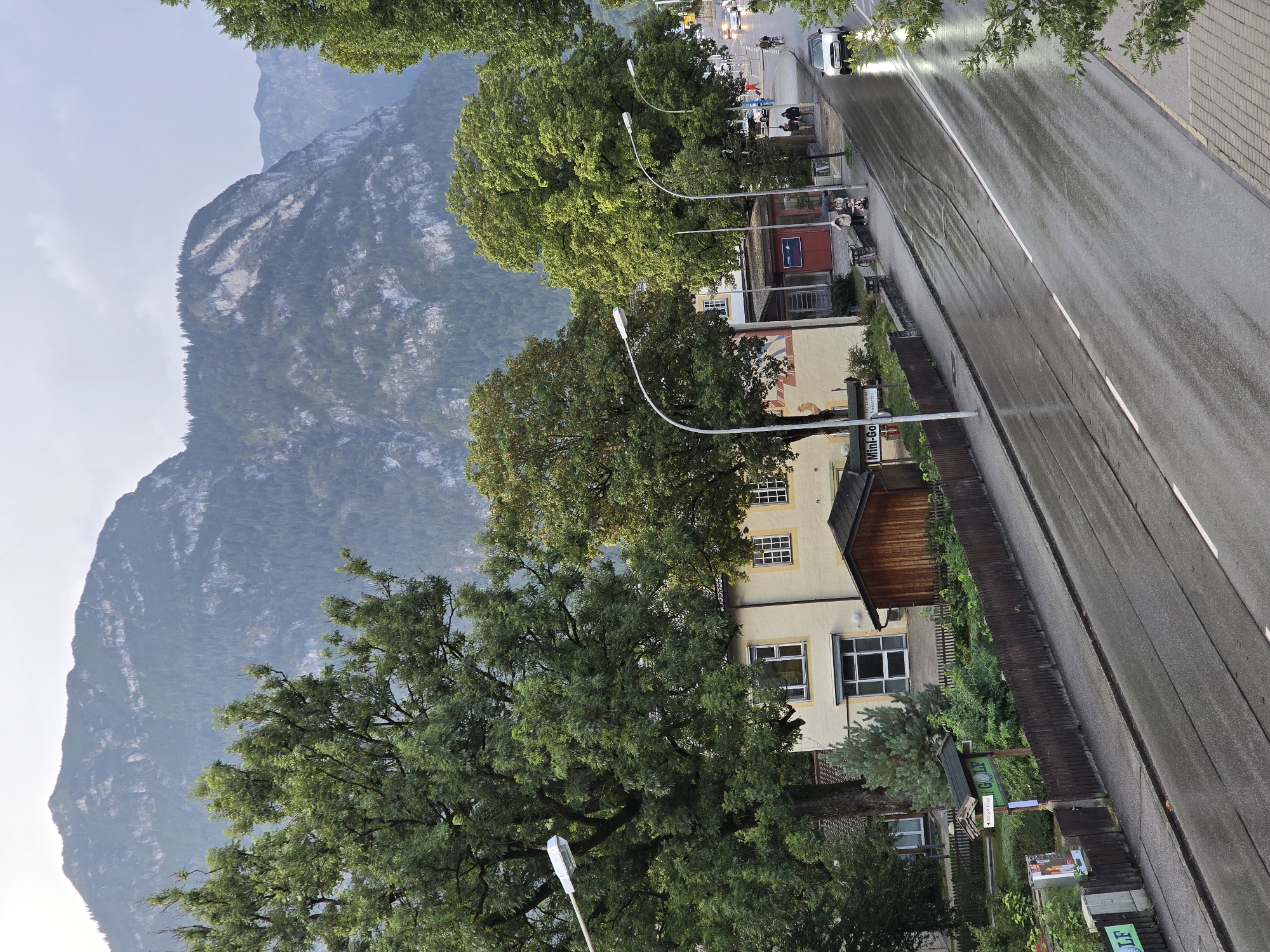 The image depicts a street scene in Garmisch-Partenkirchen, Germany, under overcast skies. A wet asphalt road dominates the foreground, reflecting the grey light. On the left side of the road, a light-colored building with multiple windows is visible, partially obscured by a large, mature tree. Adjacent to this building is a smaller wooden structure. A dark wooden fence runs along the edge of the property. Several streetlights are positioned along the road. Further down the street, more buildings and trees are present. A few pedestrians can be seen walking, and a vehicle is driving on the wet road. In the background, a majestic mountain range with forested slopes rises under a cloudy sky. The overall impression is one of a calm, slightly damp day in a scenic, mountainous town.