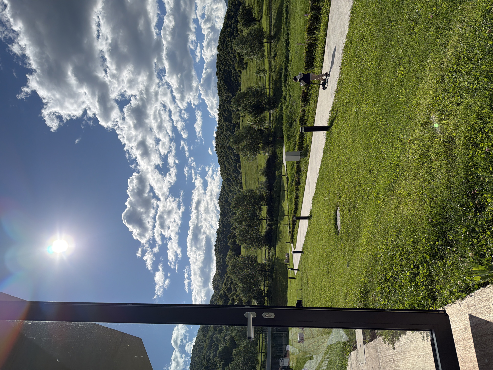 A slightly rotated, outdoor scene shows a bright sunny day with a clear, blue sky and fluffy white clouds over a grassy, green landscape with trees and a hill in the distance. The sun is bright, creating a lens flare in the upper left. A paved path runs along the edge of the grass, and a person on a scooter is visible on the path. There are short, black posts lining the path. The foreground shows part of a building, possibly a window frame or a door frame, with a handle. The scene appears to be in Chkalov, Armenia, judging by the landscape and architecture.