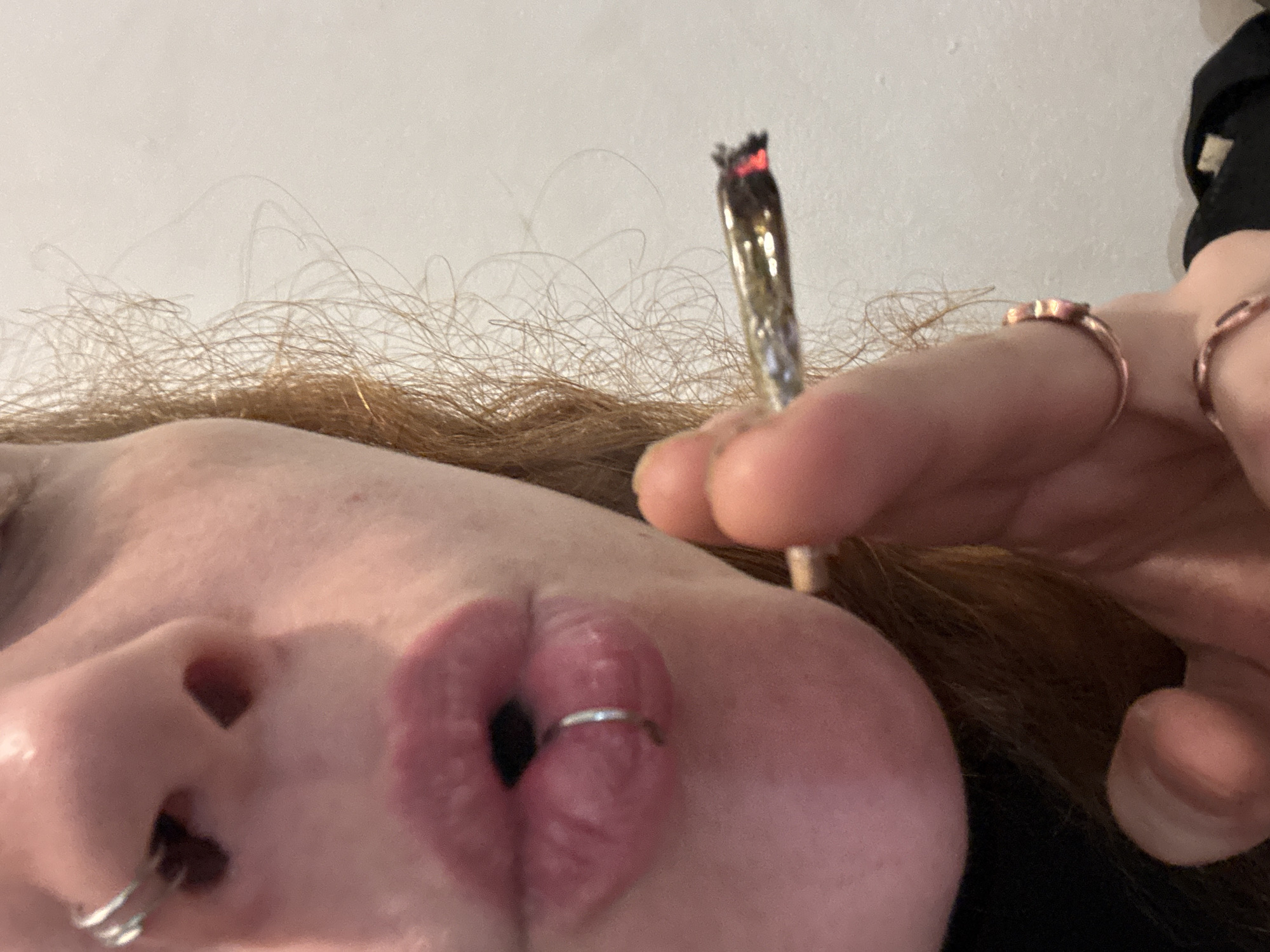 A close-up, low-angle shot captures the left side of a person's face, including their lips, nose, and fingers, against a plain, light-colored background. The individual has reddish-brown hair, visible along the top and left edges of the frame. Their lips are pursed into an O-shape, with a silver ring piercing the lower lip. Another silver ring is visible in the left nostril.
Two fingers, adorned with multiple rings, are shown holding a lit joint or cigarette. The tip of the joint is glowing brightly, indicating it is actively burning. A dark sleeve is partially visible on the right side of the image. The scene suggests the person is in the act of smoking. The setting appears to be indoors. No text is visible. This image was captured in Buenos Aires, Argentina.