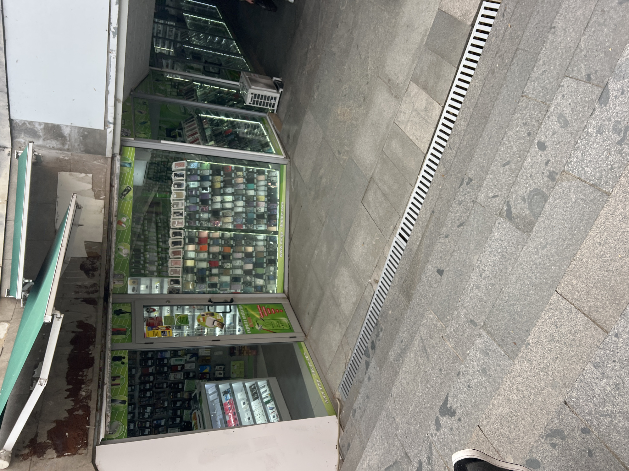 The image depicts the exterior of a shop in Plovdiv, Bulgaria. The shopfront features large glass windows displaying a variety of items, including phone cases. The shop has a green awning. The sidewalk in front of the shop is paved with grey stone tiles and includes a drainage grate. A partially visible shoe of a person is in the foreground.