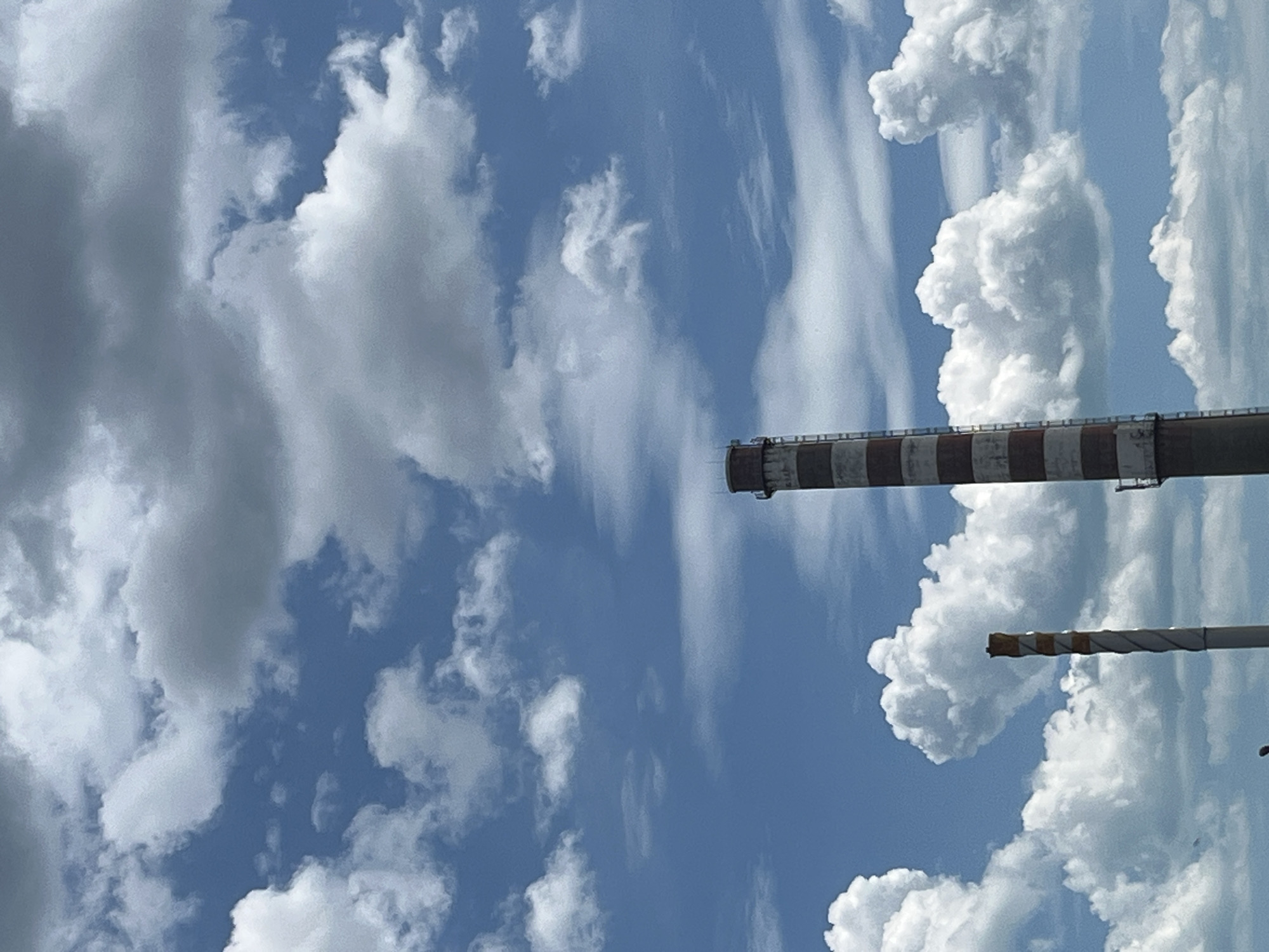 The image shows a partly cloudy sky over Varna, Bulgaria, with two industrial chimneys in the foreground. The higher chimney is marked with alternating red and white bands, while the lower chimney has yellow and white bands. The sky is blue with scattered white clouds.