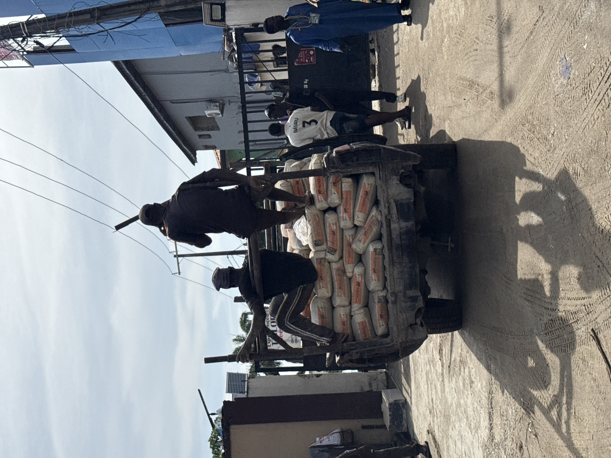 In Ado, Nigeria, a truck laden with bags of cement is being unloaded by several men. Two men are perched on the truck, handling the bags. One man stands on the ground near the truck, and two additional men are standing nearby. The surroundings include buildings, utility poles with wires, and a concrete surface. The overall environment suggests an urban setting.