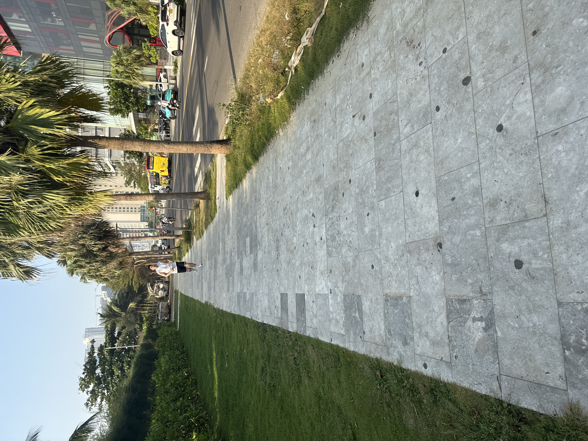 A sidewalk made of gray rectangular stones runs along a street lined with palm trees in Đà Nẵng, Vietnam. The sidewalk is bordered by green grass. A person in a white shirt and dark shorts is walking along the sidewalk. The street is parallel to the sidewalk and has cars and motorbikes. Buildings can be seen in the distance. The sky is light blue.