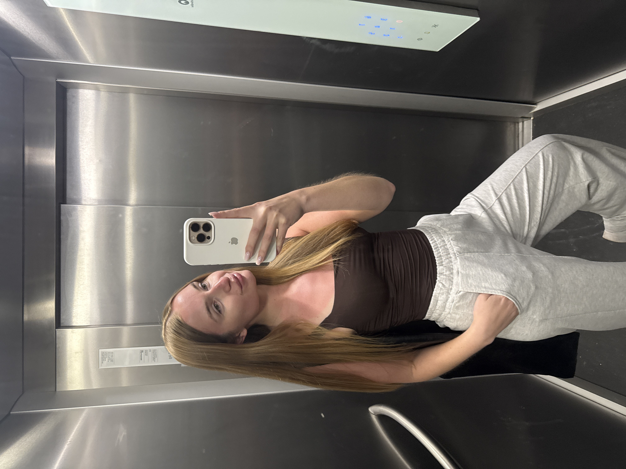 A young woman is captured in a selfie taken inside a modern elevator, located in Budva, Montenegro. The image is rotated 90 degrees clockwise, depicting her as if leaning against the elevator wall. She has long, straight light brown hair and a fair complexion. Her attire consists of a dark brown strapless top and light grey sweatpants, with one hand casually placed in a pocket. She holds a white smartphone, likely an iPhone with a triple-lens camera, with the other hand to take the picture; her nails are manicured with a light, natural polish.
The elevator's interior features brushed stainless steel walls and a dark floor. A modern control panel is prominently visible in the upper part of the image, displaying a digital "0" and several blue illuminated buttons indicating floor numbers and other functions. The panel shows some reflections and smudges. A smaller silver plaque with unreadable text is mounted on the left wall. The woman maintains a neutral and composed expression, characteristic of a casual selfie. The setting suggests a contemporary indoor environment.
