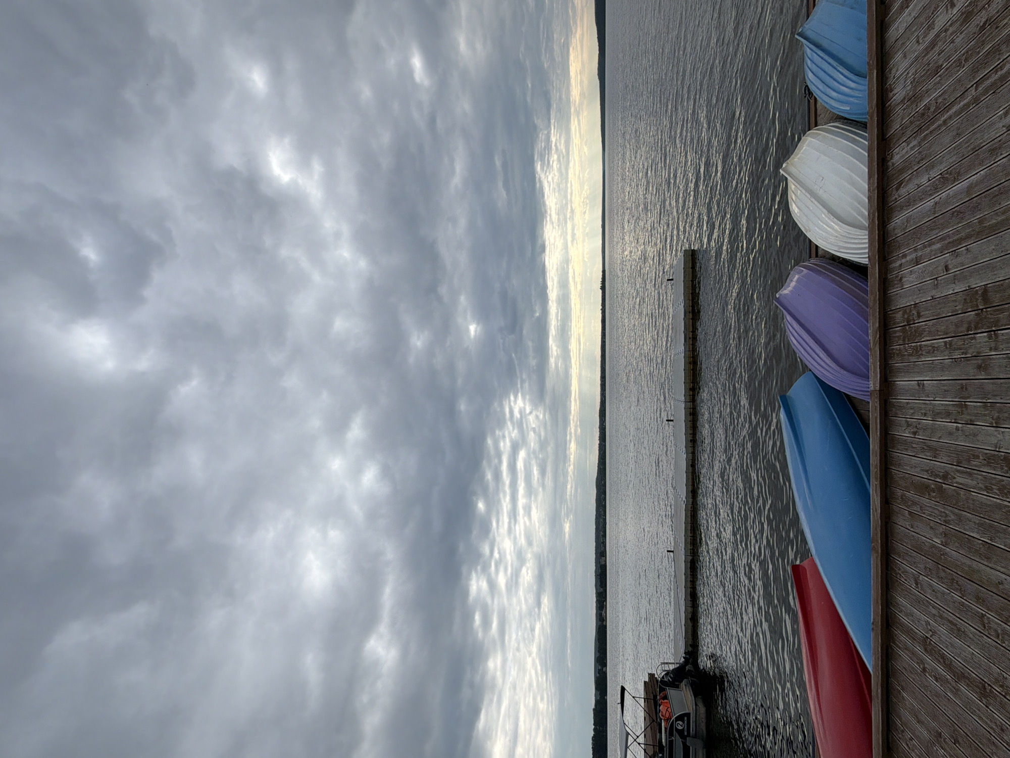 The image depicts a cloudy day over a tranquil lake in Votkinsk, Russia. Several small, colorful rowboats are lined up on a wooden dock in the foreground. The boats are inverted, with their hulls facing upwards, displaying vibrant red, blue, purple, and white colors. In the background, a floating dock extends into the calm water, and a boat is moored to it. The horizon is visible, with a tree-lined shore in the distance. The sky is filled with a dense layer of gray clouds, with some hints of sunlight breaking through. The overall atmosphere is serene and slightly overcast.