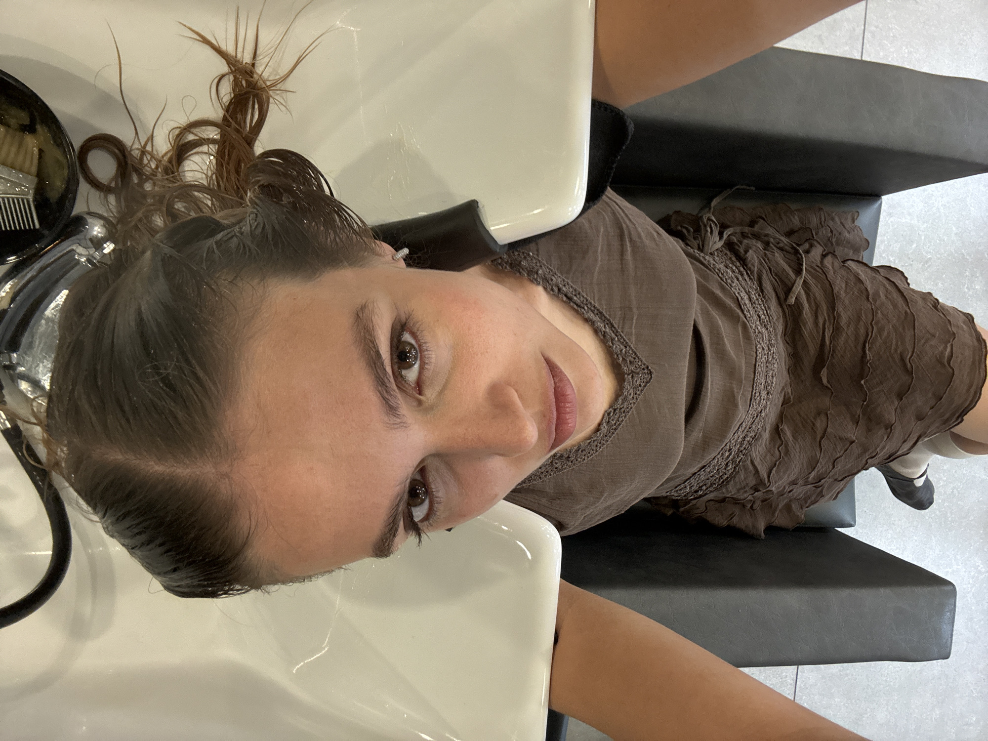A woman lies reclined at a hair salon sink in Tbilisi, Georgia. Her damp brown hair partially rests in the white basin. She has brown eyes and pink lips, and wears a brown dress with ruffled details. A black salon chair is visible, and salon implements are present in a container to the left. The floor is light-colored tile.