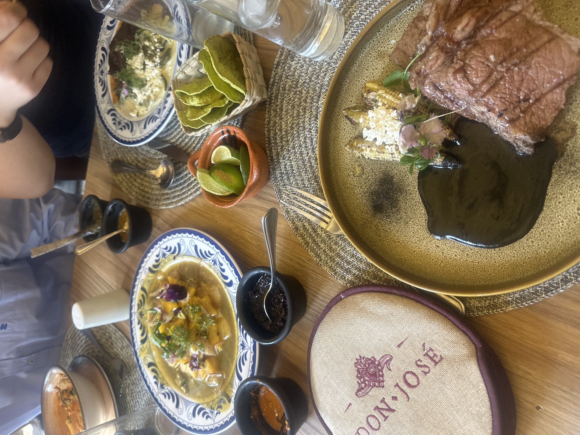 The image shows a table at a restaurant in San Martín de las Pirámides, Mexico, featuring a variety of dishes. A partially visible person is seated at the table. The food includes a steak served on a dark sauce with what appears to be baby corn and cheese. Additional dishes are served in bowls with blue and white patterned rims. One bowl contains a soupy dish with vegetable matter and flowers; another small bowl contains black sauce with a spoon. There are also green tortilla chips in a small basket and a small clay pot containing lime wedges. The table is covered with a woven placemat, and there are drinking glasses, a coaster, and additional sauces. The scene suggests a shared meal at a traditional Mexican restaurant.