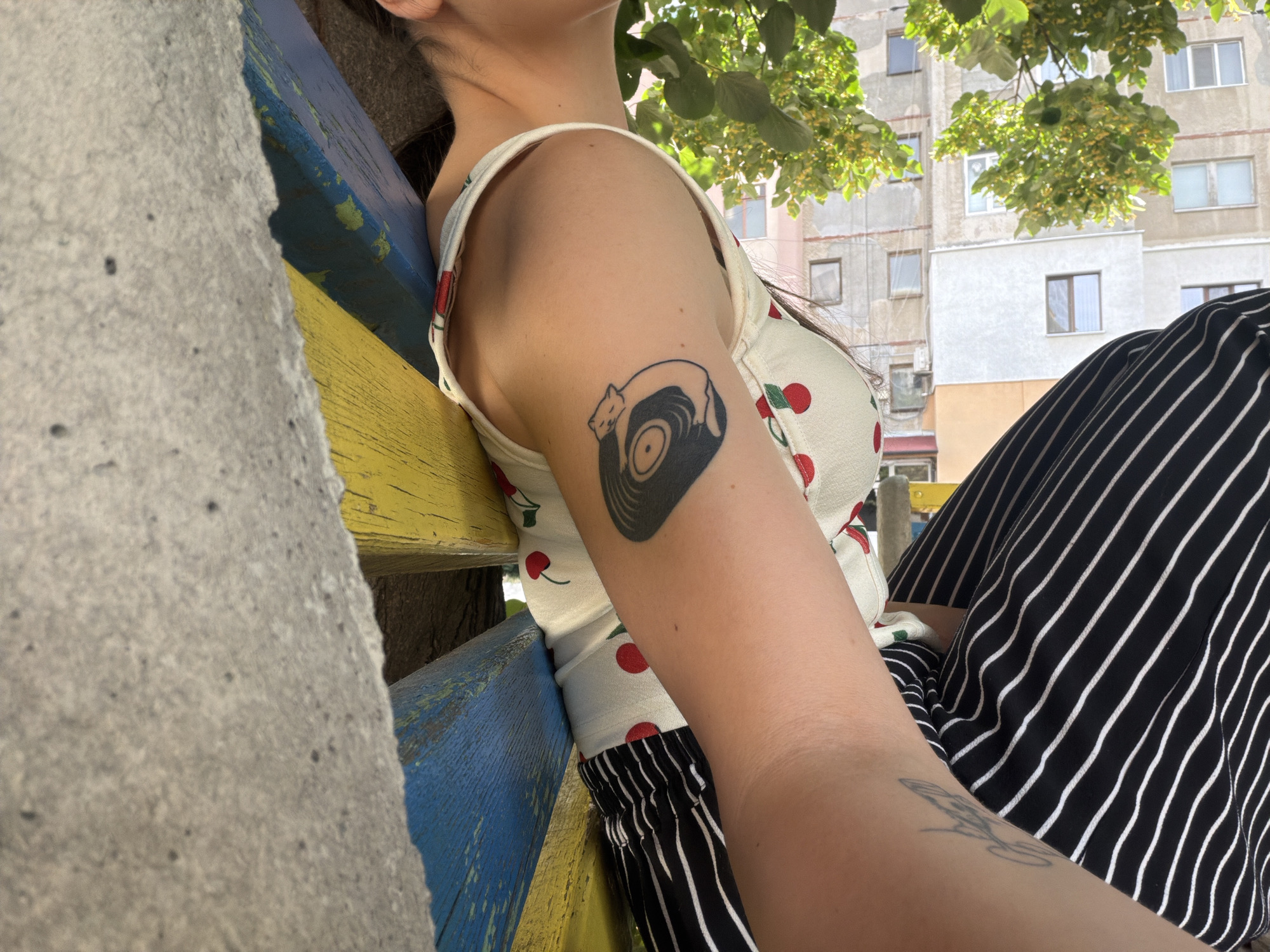 A person is sitting on a bench. The bench is painted in blue and yellow. The person is wearing a white top with red cherries and a black and white striped skirt. The person has tattoos on their arm. The tattoo on their upper arm is a black vinyl record with a cat lying on top of it. The tattoo on their forearm is a cursive design. The background shows a building and trees. The time of day appears to be daytime. The weather appears to be sunny.