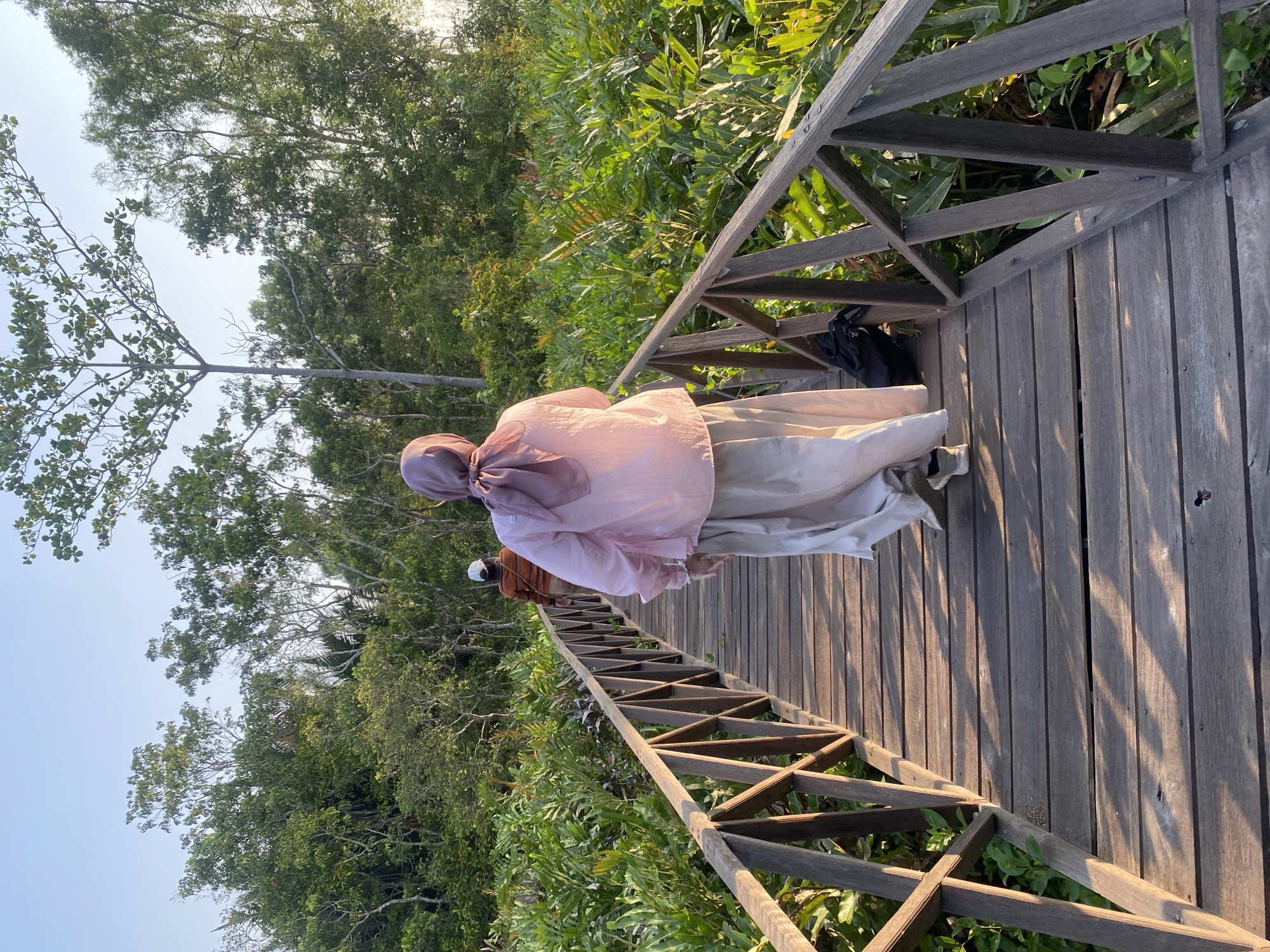 The image shows a wooden boardwalk winding through lush green vegetation. Two individuals are visible on the boardwalk. The person in the foreground, from the back, is wearing a pink headscarf and a pale pink long-sleeved shirt over a long, light-colored skirt. They are wearing white and grey sneakers. A black backpack rests on the boardwalk behind them. Another individual, further ahead and also seen from behind, is wearing a striped long-sleeved shirt and a light-colored cap. The setting appears to be a natural environment, possibly a park or conservation area, with dense foliage on either side of the boardwalk. The lighting suggests it is either early morning or late afternoon, with the sun casting long shadows across the wooden planks. The location is identified as Sungai Pitung, Indonesia.