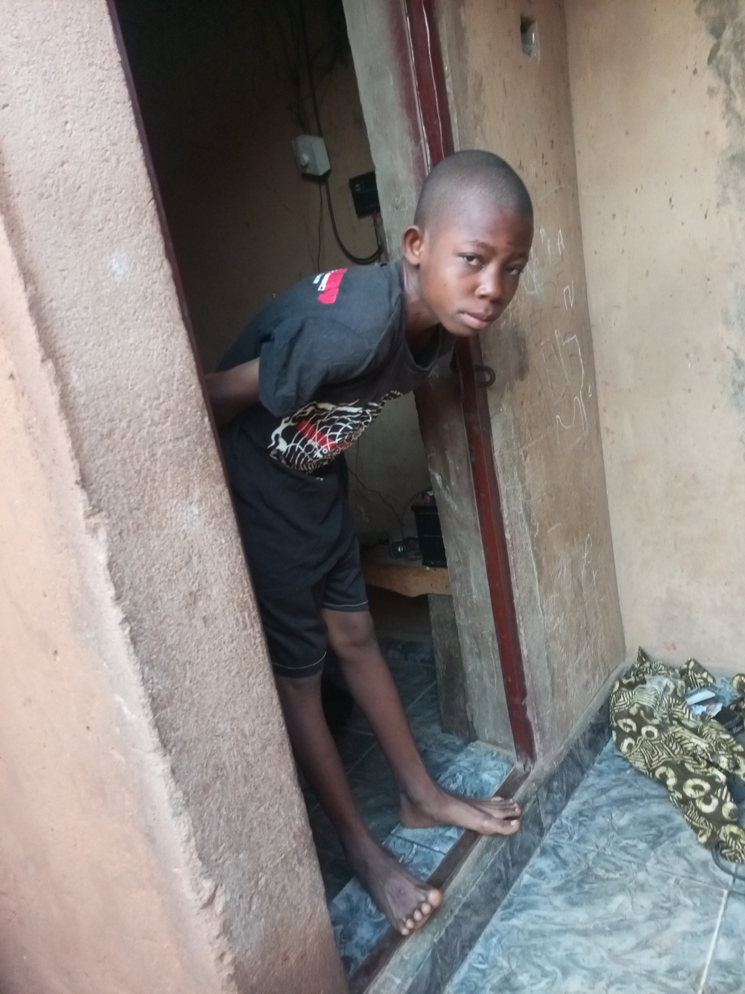 A young Black male, appearing to be of school age, stands in a doorway. He is wearing a dark t-shirt with a graphic design and dark shorts. His posture suggests he is leaning forward and looking at the camera. The environment appears to be an indoor or semi-outdoor setting, possibly a residential building, with textured walls. One wall to the right of the doorway has some faint white markings on it. On the floor in the foreground, there are tiled surfaces and a pile of patterned fabric. To the left of the doorway, a portion of a textured, light-colored wall is visible. The lighting suggests it is daytime, with light coming from the direction of the camera. There are no overt signs of distress or danger. The image provides a close-up view of the boy and his immediate surroundings.
