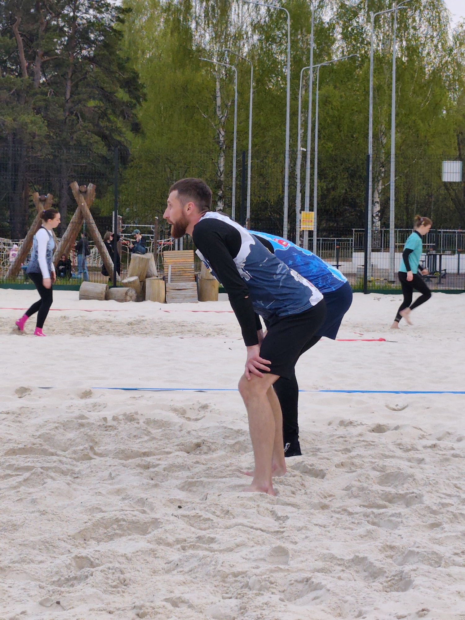 The image captures a scene on a beach volleyball court located in the city of Chaikovsky, Russia. Two men are standing on the sandy court, both barefoot and leaning forward with their hands on their knees, seemingly in a ready or resting position. They are dressed in athletic attire consisting of long-sleeved shirts and shorts.
In the background, a woman wearing a long-sleeved white and blue top, black leggings, and pink shoes walks near a small wooden playground structure. Another woman wearing a light green long-sleeved shirt and black leggings is walking barefoot on the far right of the frame. A blue line is visible on the sand, marking the boundary of the court.
Behind the court, a fence is visible, along with several tall, silver lampposts. The background also features trees, including birch trees, and glimpses of a park-like setting. The sky is overcast, suggesting it might be a cloudy day.